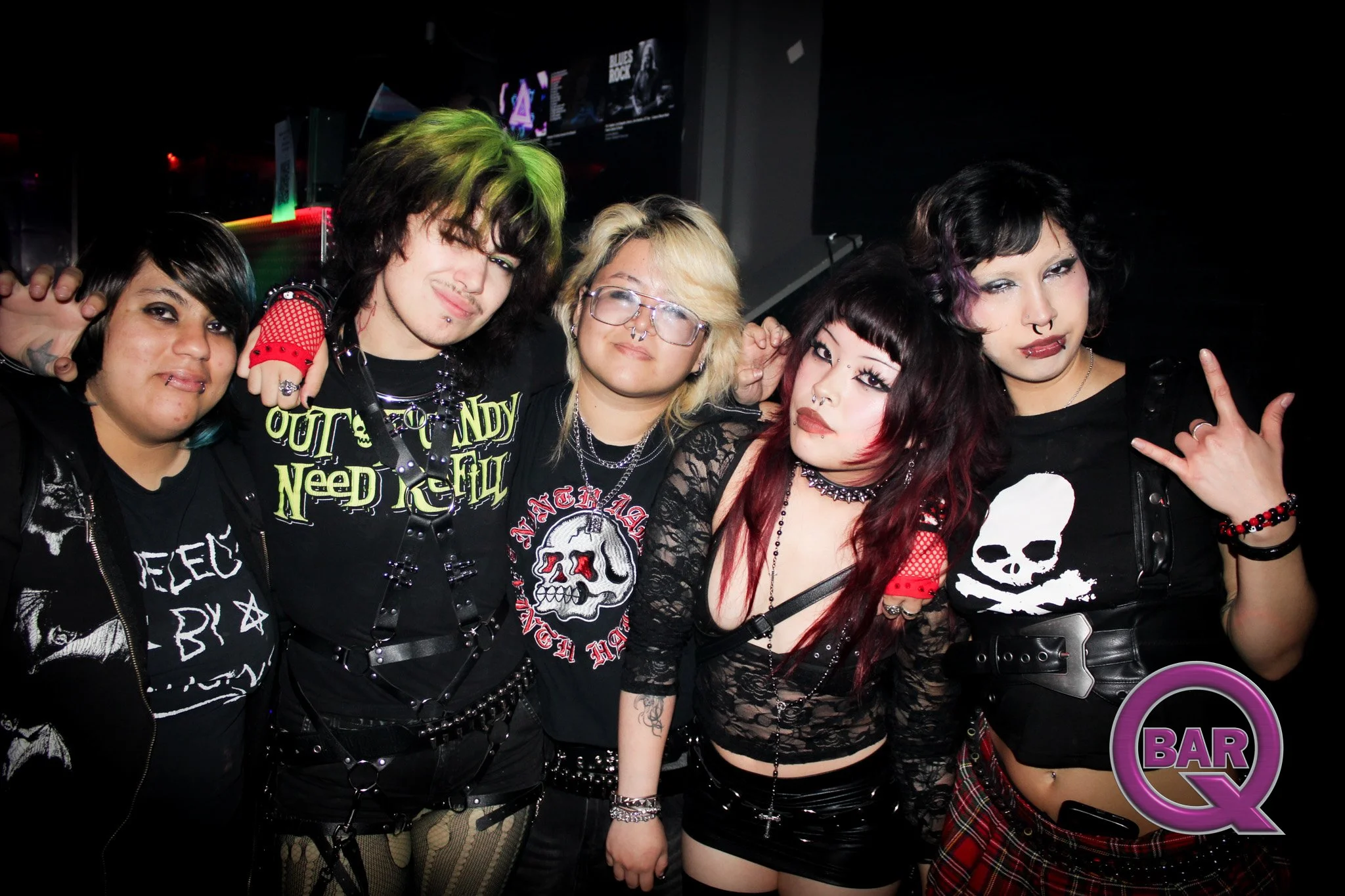 Group of five young women in punk and goth fashion at a bar or nightclub, with dark hair, colorful hair, tattoos, makeup, and edgy jewelry, posing together for a photo.