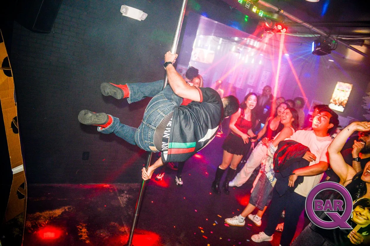 A man performing a pole dance on stage at a nightclub with a crowd of young women cheering and enjoying the performance under colorful lighting.