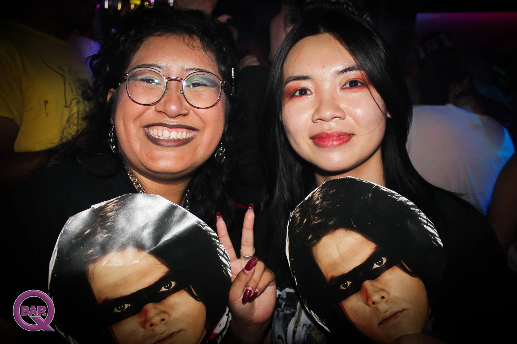 Two women smiling at a nighttime event, holding posters of a person's face, with somber makeup, dark hair, and intense eyes. One woman is wearing glasses and dark lipstick, while the other has a subtle makeup look. They are close together, with one w