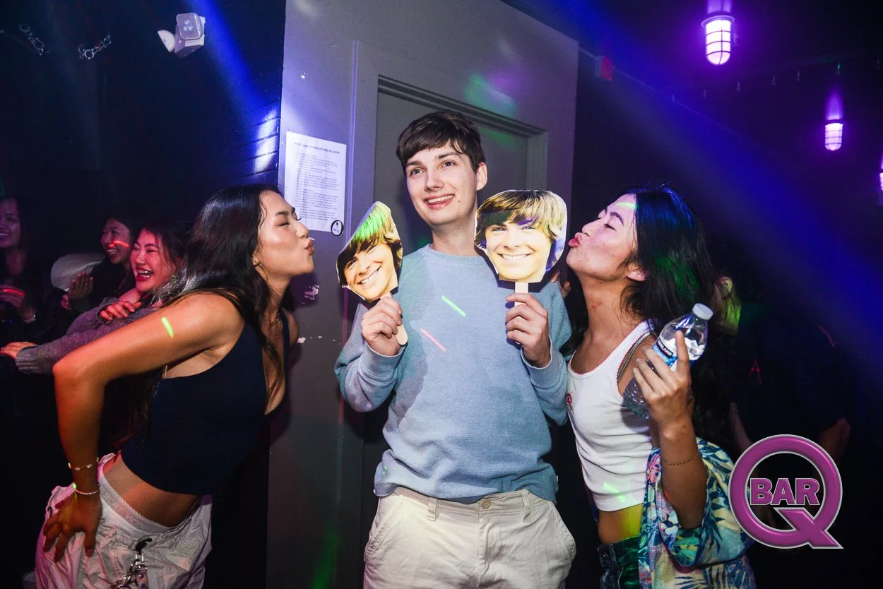 People at a party holding photo masks of a young man's face, smiling, with colorful lights in the background, and a ring light on the ceiling. Two women are puckering their lips toward the man, who is smiling and holding two cutouts of the same face.