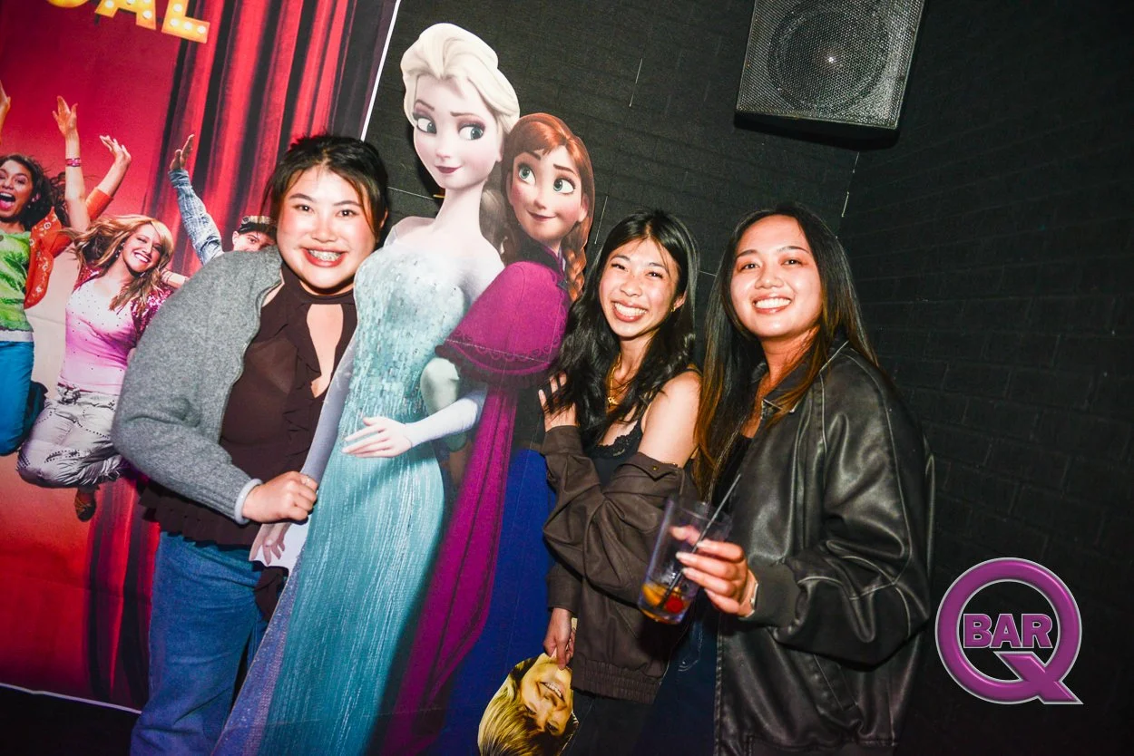 Three women smiling and posing with a life-sized cardboard standee of Disney's Elsa and Anna from Frozen at a bar or party. One woman in the middle holds a drink.