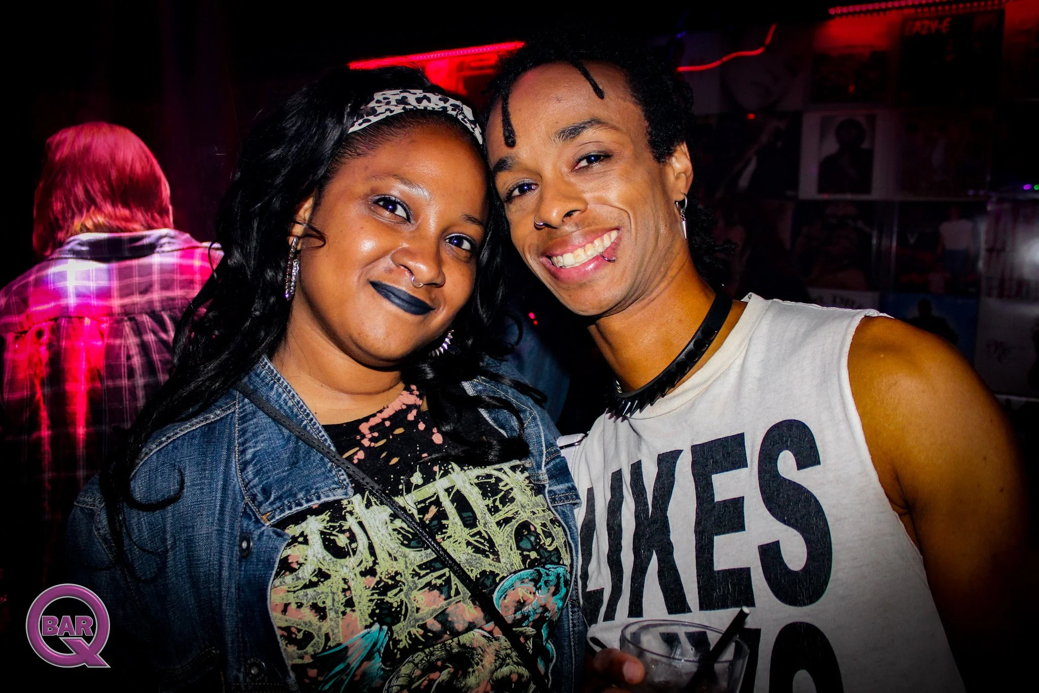 Two people smiling at a bar with red lighting; woman with dark hair, wearing a leopard print headband and dark lipstick, and man with dreadlocks, earrings, nose ring, and sleeveless shirt with text.