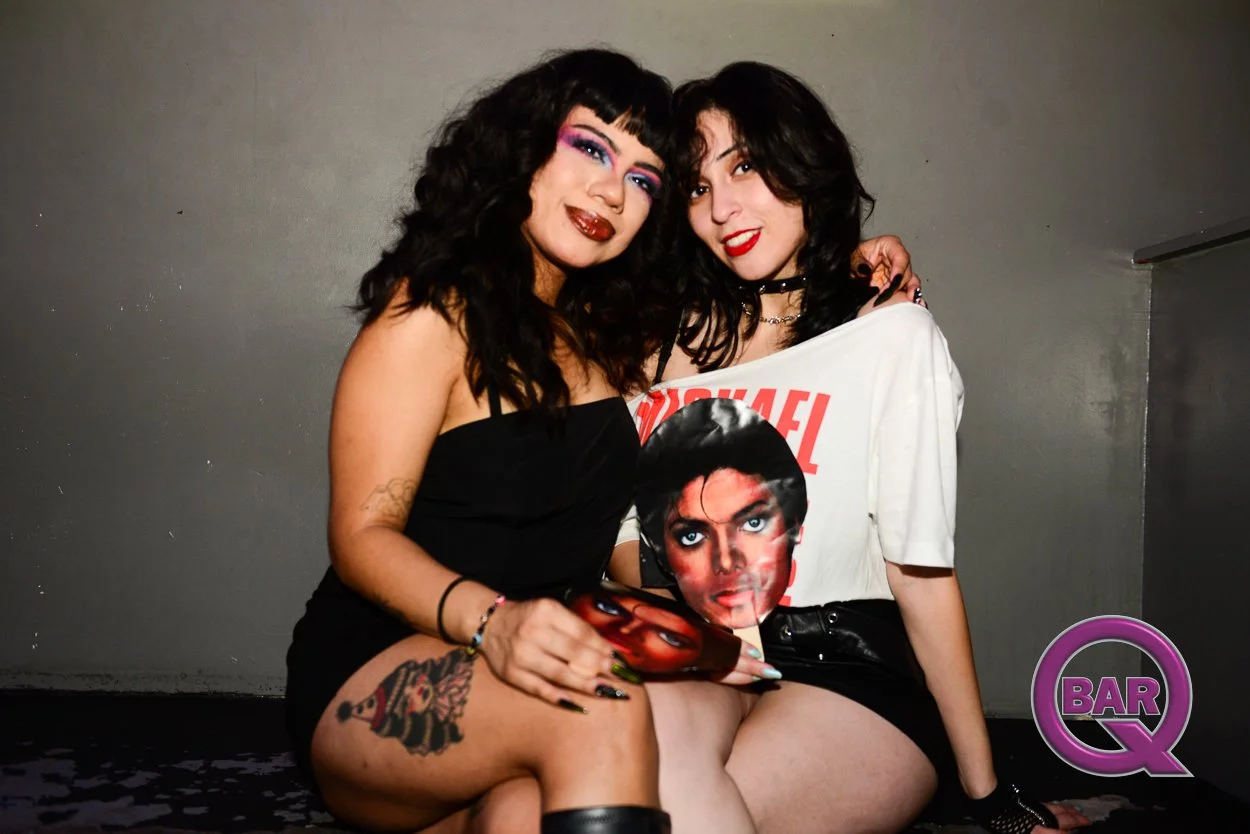 Two women with dark curly hair and makeup sitting close together, smiling at the camera. One is wearing a black strapless top, and the other is wearing a white vintage T-shirt featuring Michael Jackson. One woman has a tattoo on her thigh and holds a
