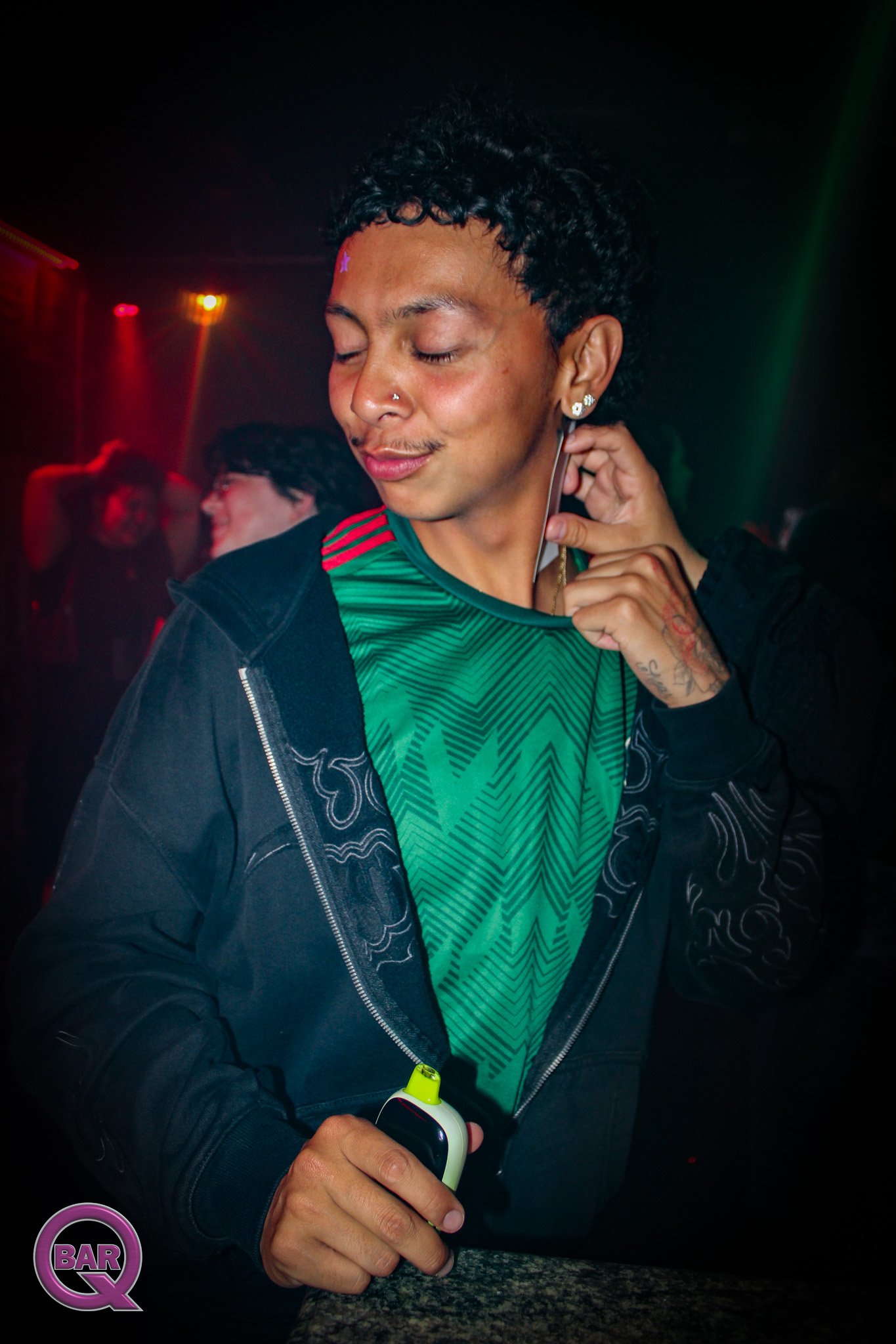 A man with a nose piercing and earrings holds a device in a dark, club-like environment with red and green lighting, next to a table with a bar logo.