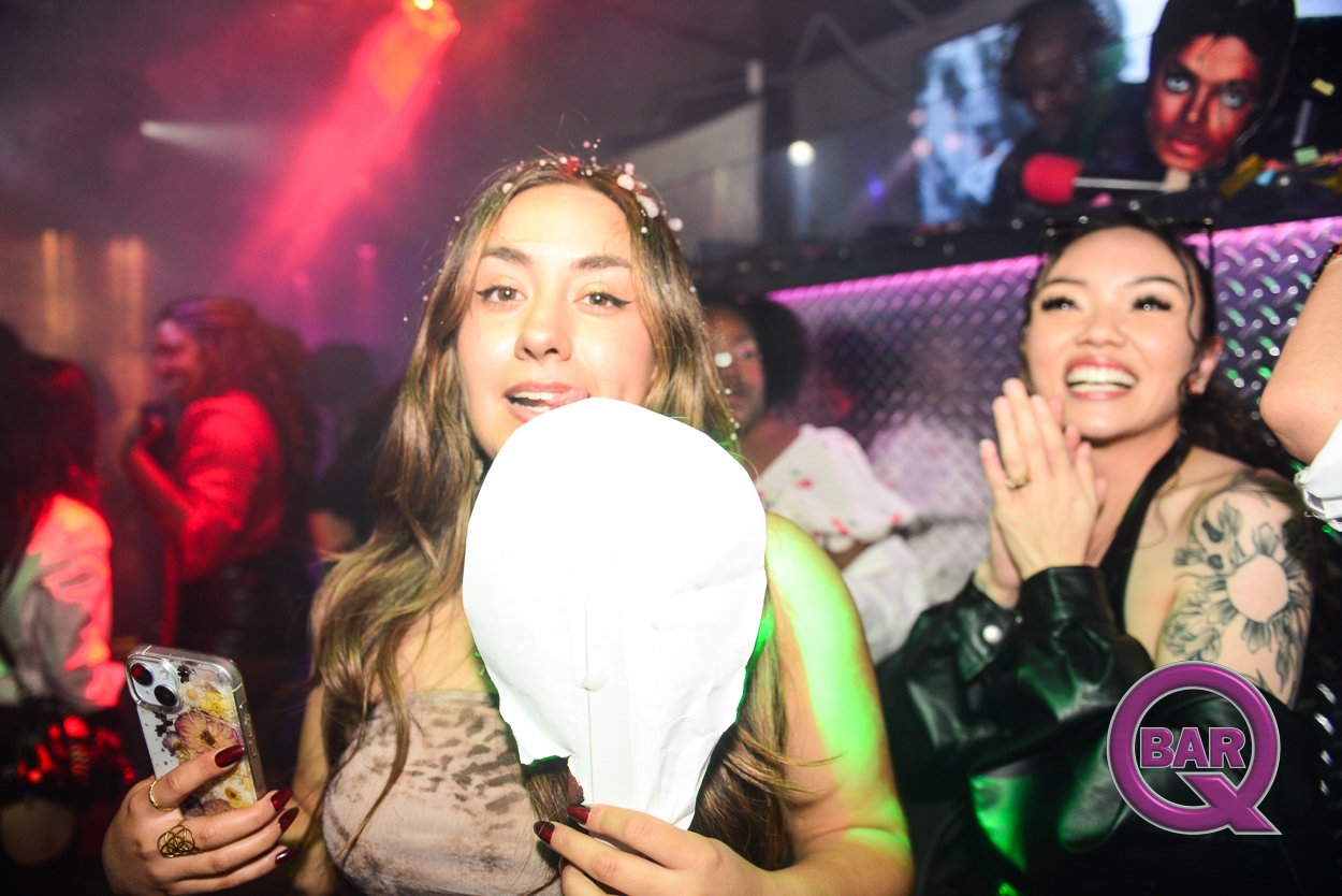 Group of women enjoying a party at a nightclub, with one woman holding a paper lantern and a smartphone, smiling and clapping, with colorful lighting and a bar logo in the corner.