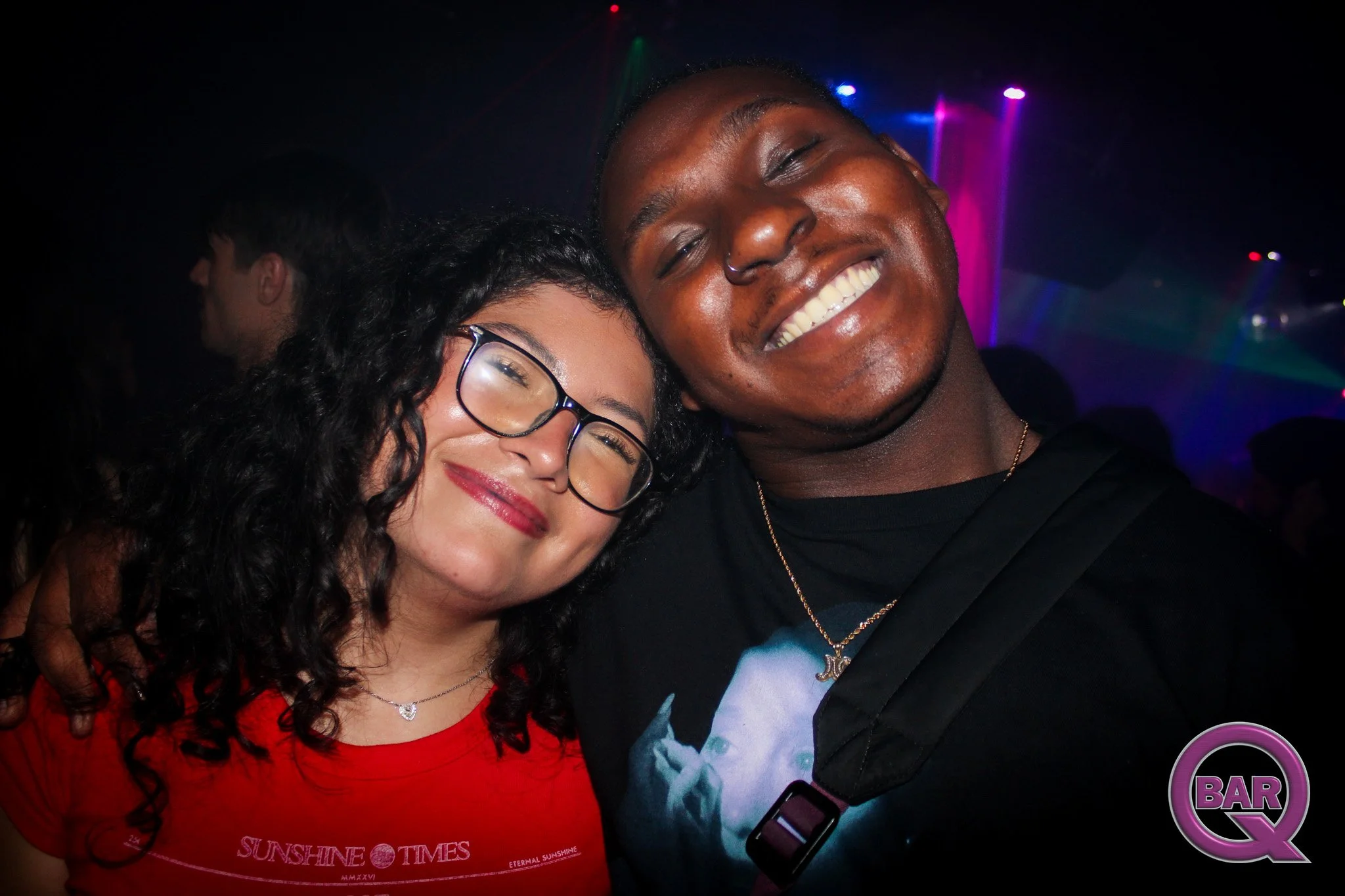 Two people smiling and hugging at a nightclub or bar with colorful lights in the background.
