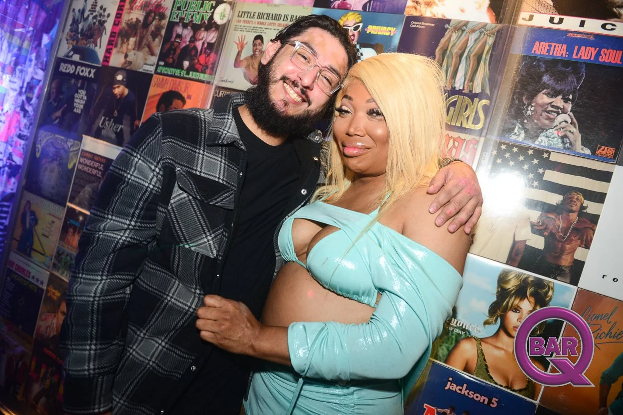 Two people, a man with glasses and a woman with blonde hair, pose together in front of a wall covered with colorful album covers. The woman is wearing a light blue off-shoulder crop top and matching skirt, and the man is wearing a black shirt and a p