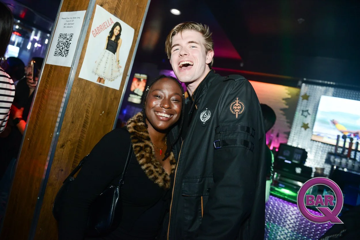 Two people smiling and hugging at a bar with colorful lights in the background. The person on the left is a woman with dark skin, wearing a black outfit with a leopard print fur collar. The person on the right is a tall young man with light skin and 