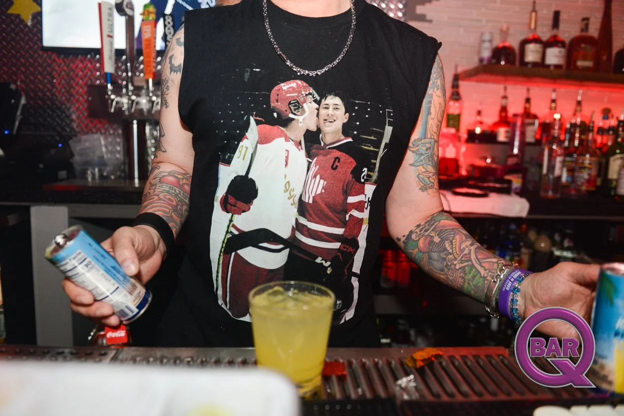 Person standing at a bar counter wearing a sleeveless black shirt with a hockey player graphic, tattoos on arms, holding a can and a yellow drink.