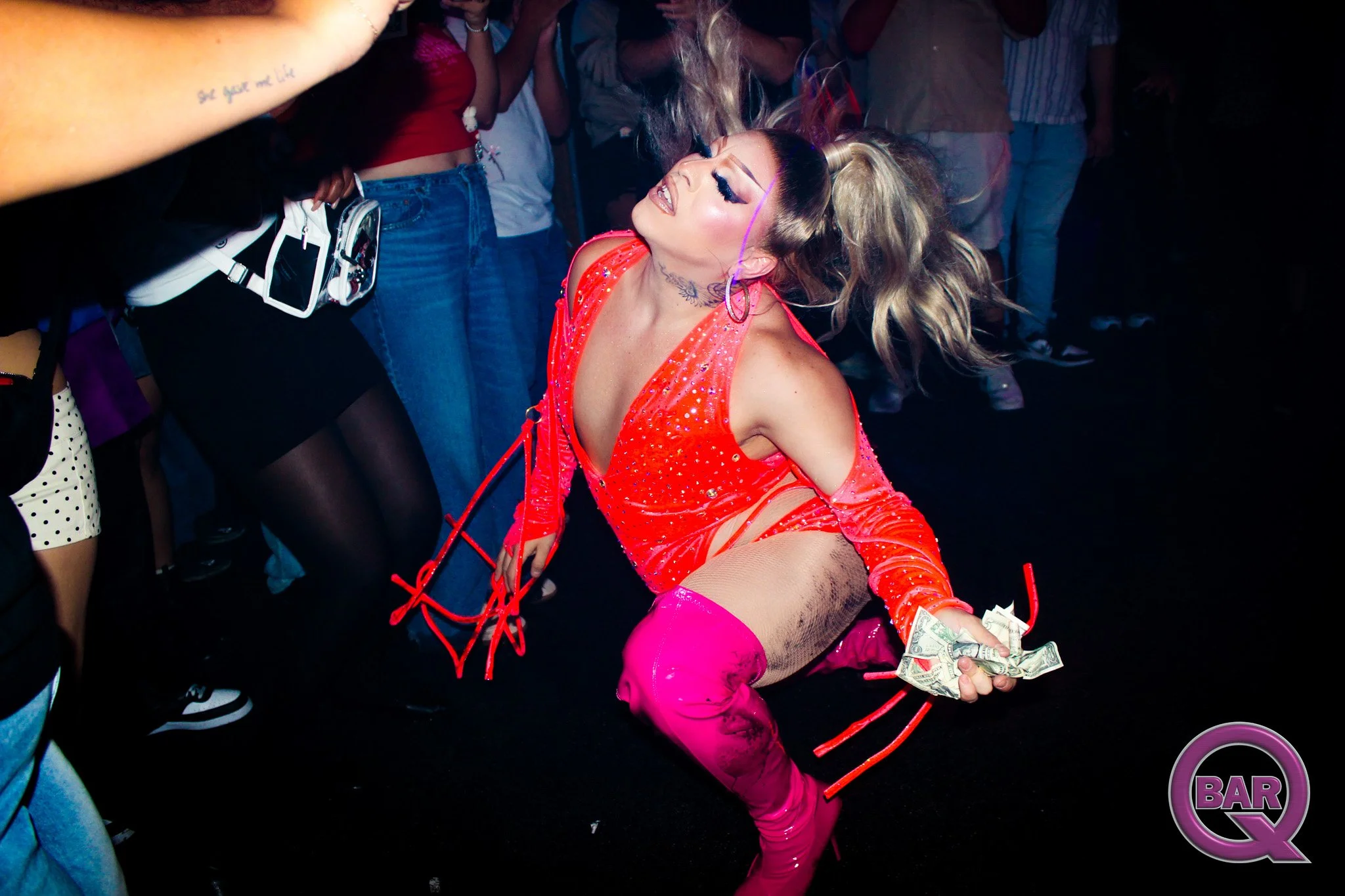 A woman with long hair styled in a high ponytail dancing in a crowded nightclub, wearing a revealing bright orange outfit with sparkles, pink thigh-high boots, and holding dollar bills. She is smiling with her eyes closed, surrounded by people.