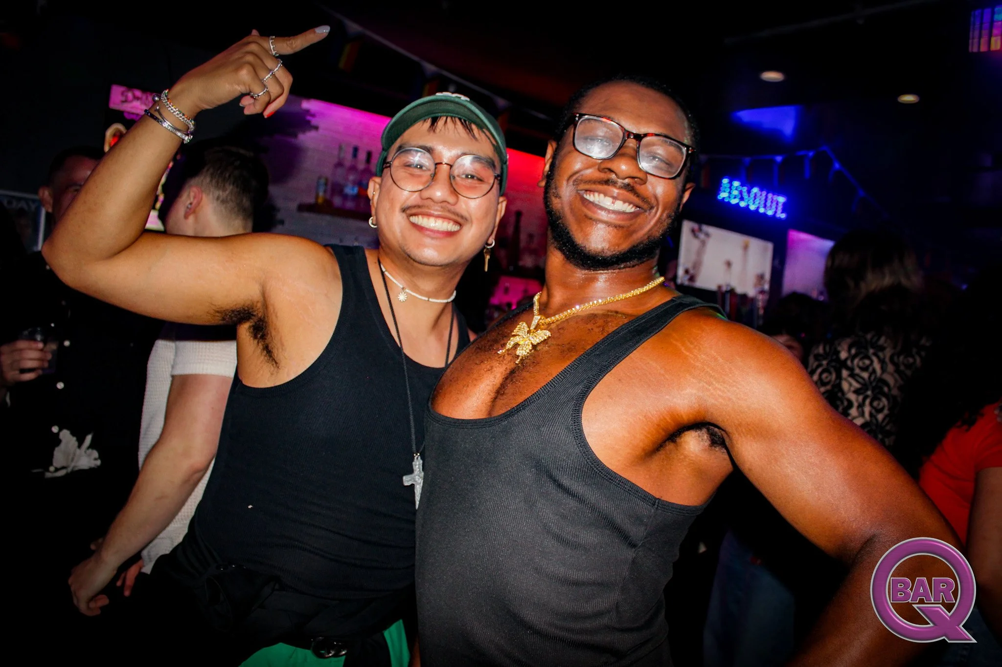 Two happy men smiling together at a bar or nightclub, wearing tank tops, with multicolored lighting in the background.