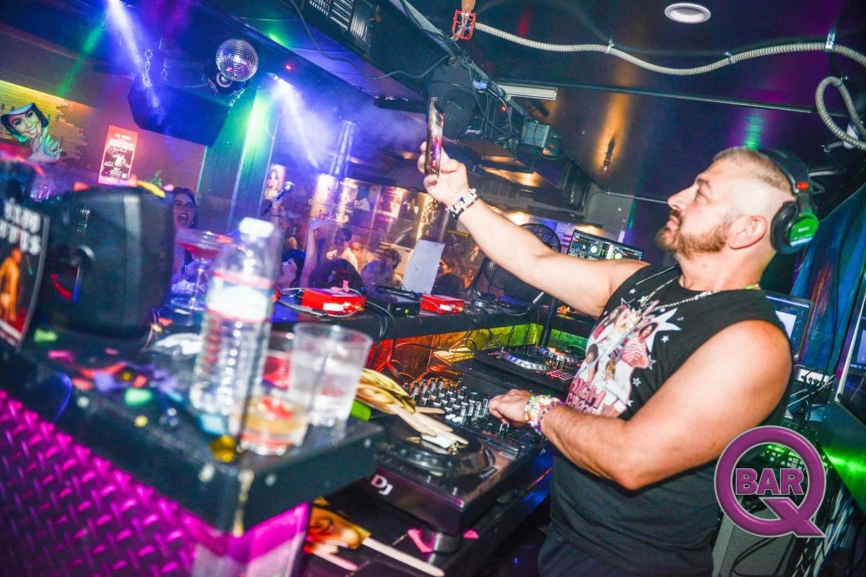 A male DJ wearing a sleeveless shirt and headphones, holding a smartphone to take a photo in a nightclub. The club has colorful lighting, a dance floor with people, and a purple 'BAR Q' logo in the bottom right corner.