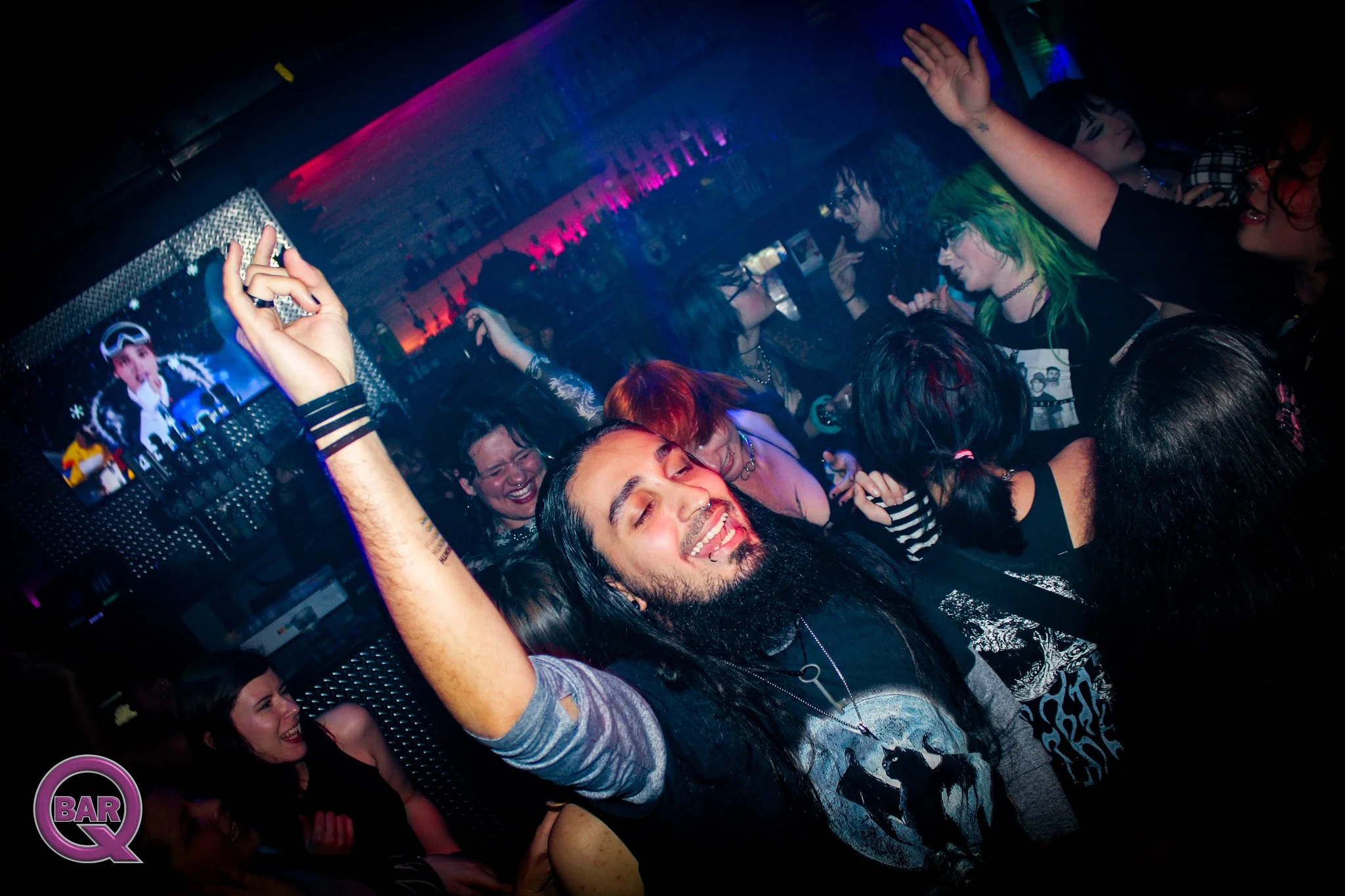 People dancing and enjoying at a nightclub with colorful lighting, accompanied by a DJ and a monitor displaying a performer, likely a singer.