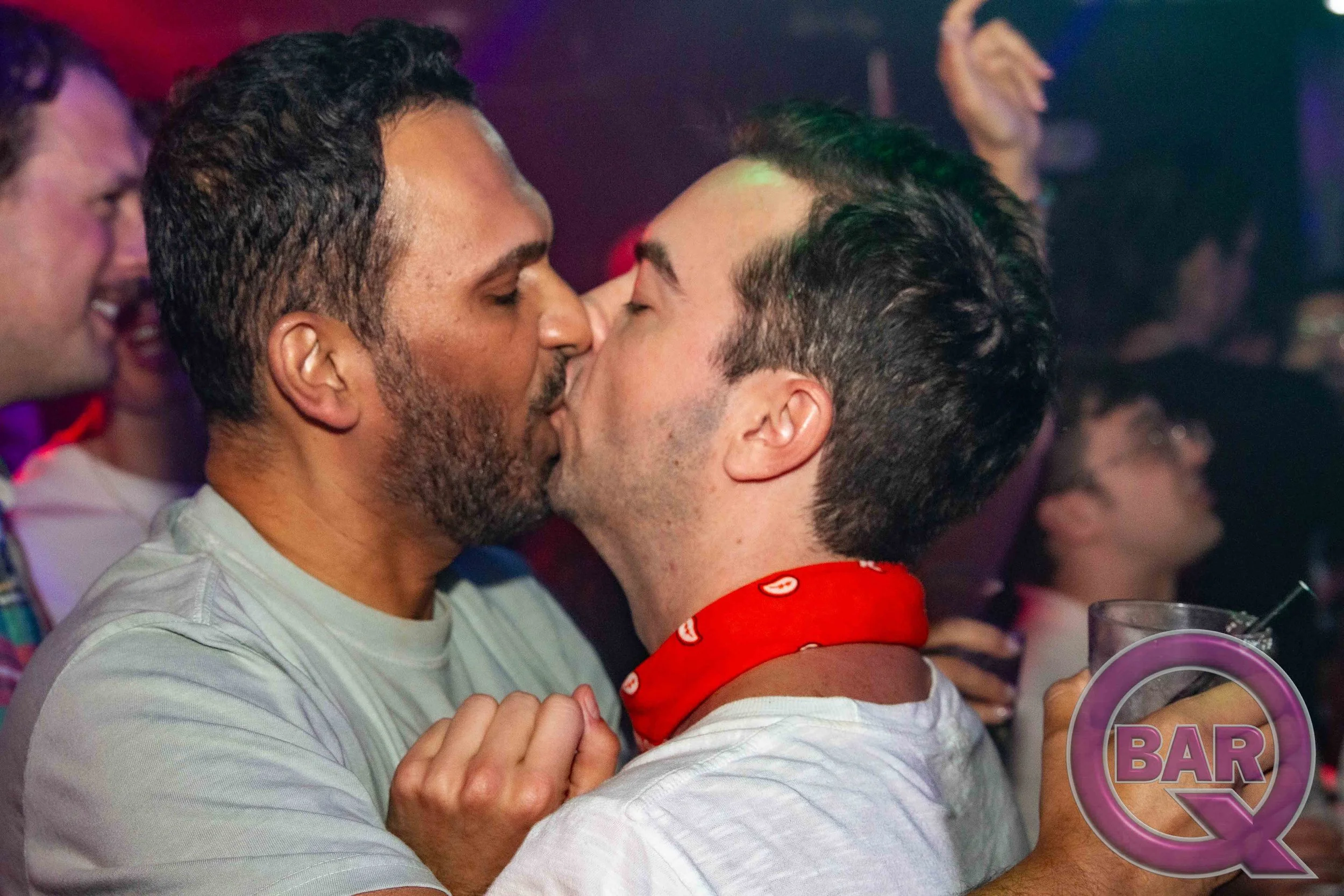 Two men with dark hair and beards are kissing at a social gathering or party, with other people visible in the background