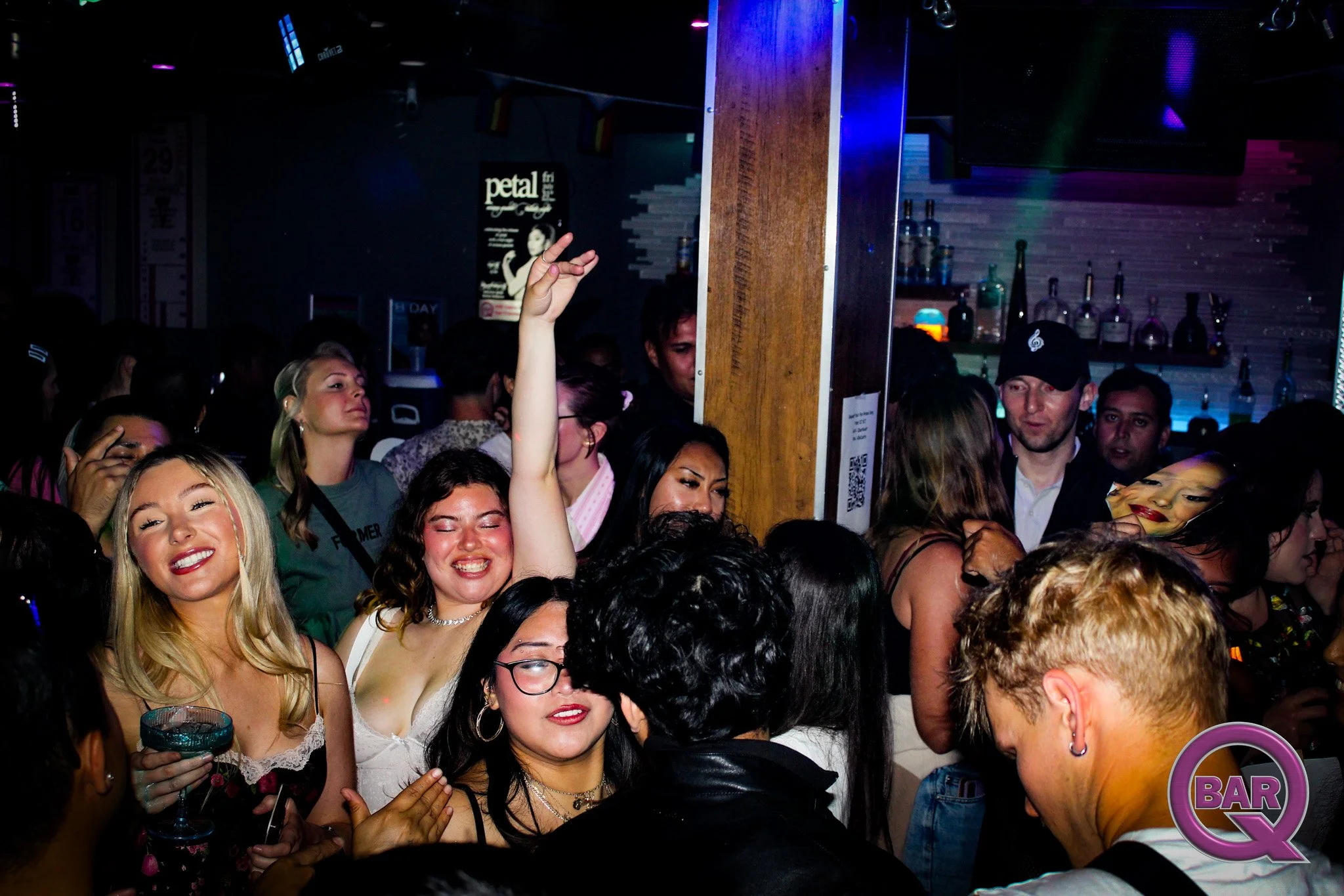 A crowded bar with people dancing and socializing, a woman in the foreground smiling and holding a drink, others raising their hands, and a bartender behind the bar with bottles and glasses, illuminated by colorful lights.