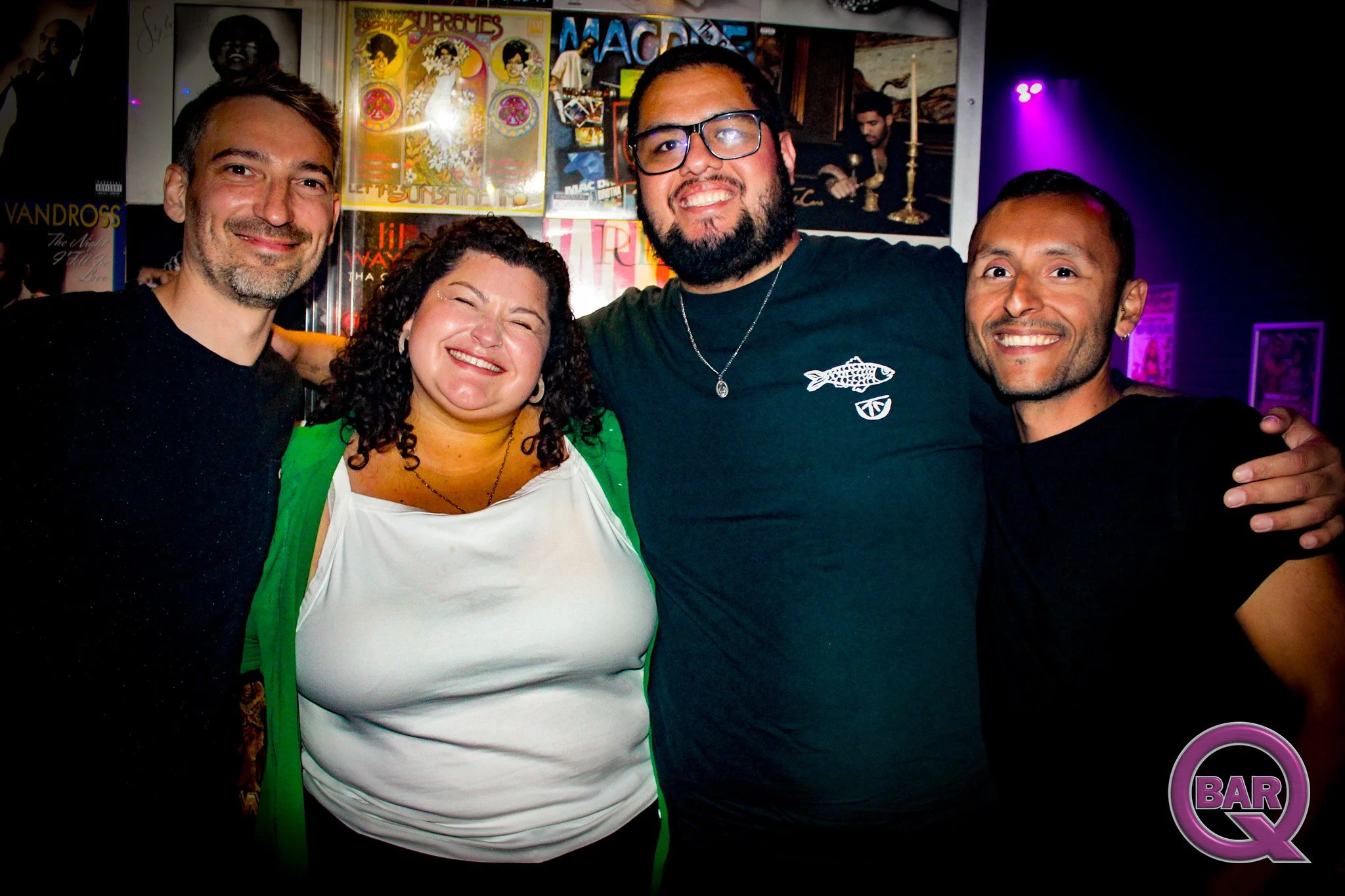 Group of four friends smiling, standing close together in a bar or club, with posters and black walls in the background.
