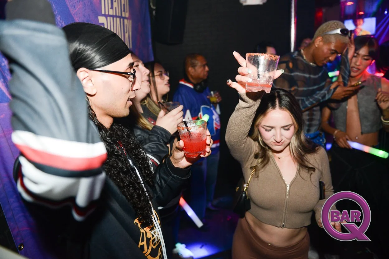 Group of people dancing and socializing at a nightclub, with colorful lighting and glow sticks, some holding drinks.