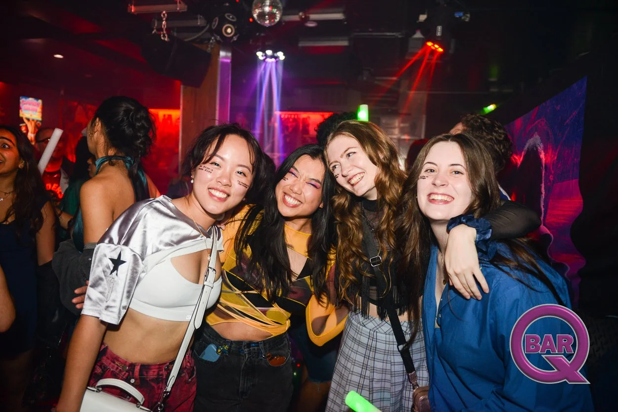 Four young women smiling and celebrating in a bar or nightclub with vibrant lighting and people in the background.