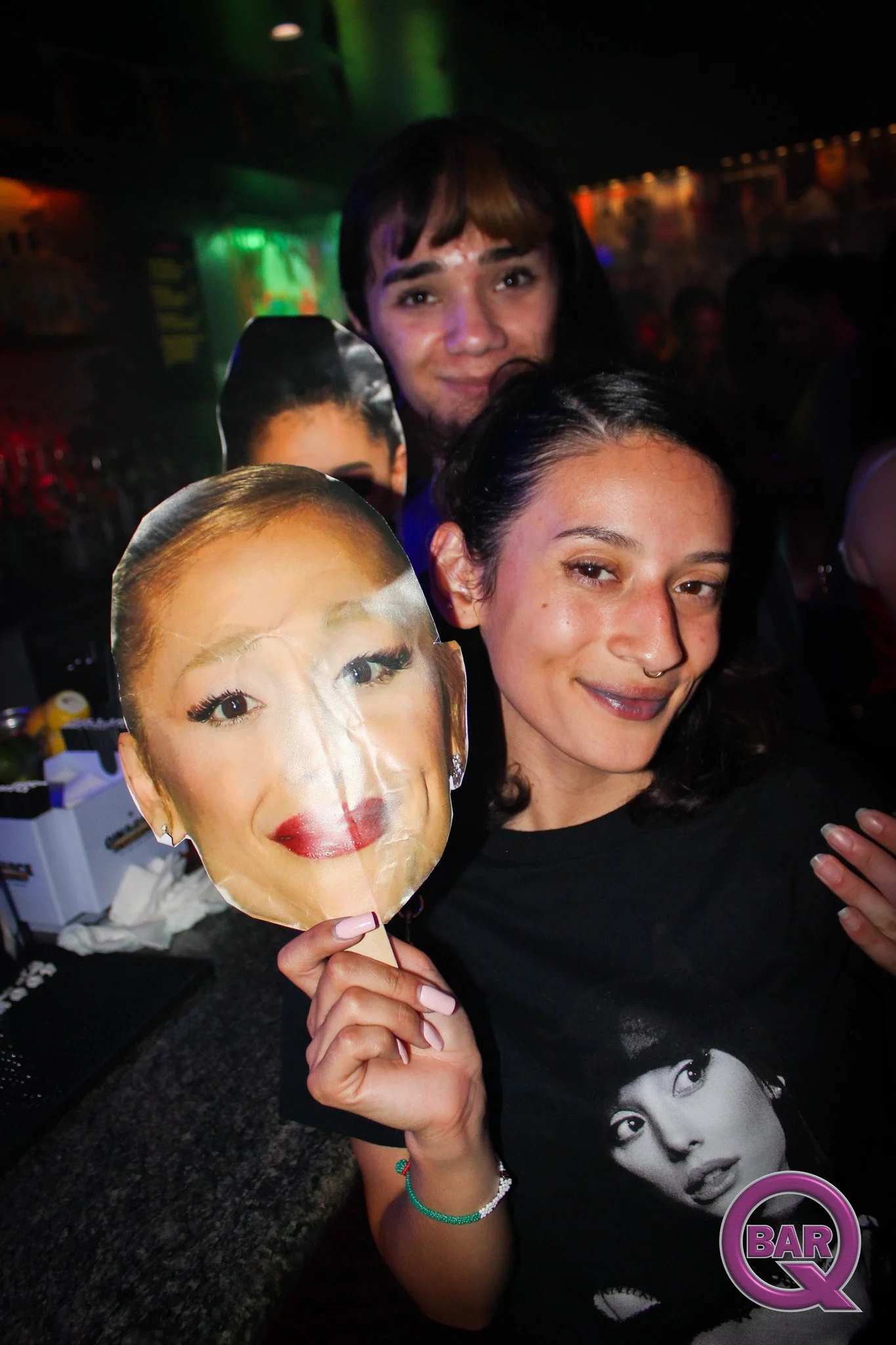 A woman with dark hair and a nose piercing holding a Marie Antoinette face mask at a bar, with other people in the background and colorful lighting.