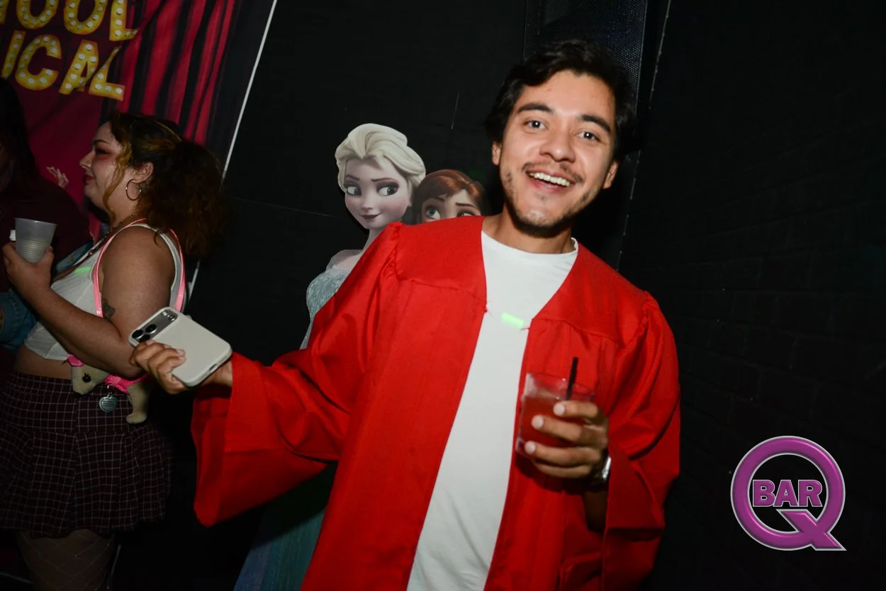 A young man wearing a red graduation gown and a white t-shirt, smiling and holding a drink and a smartphone, at a party with Disney Princess decorations, including Elsa and Anna from Frozen, in the background.