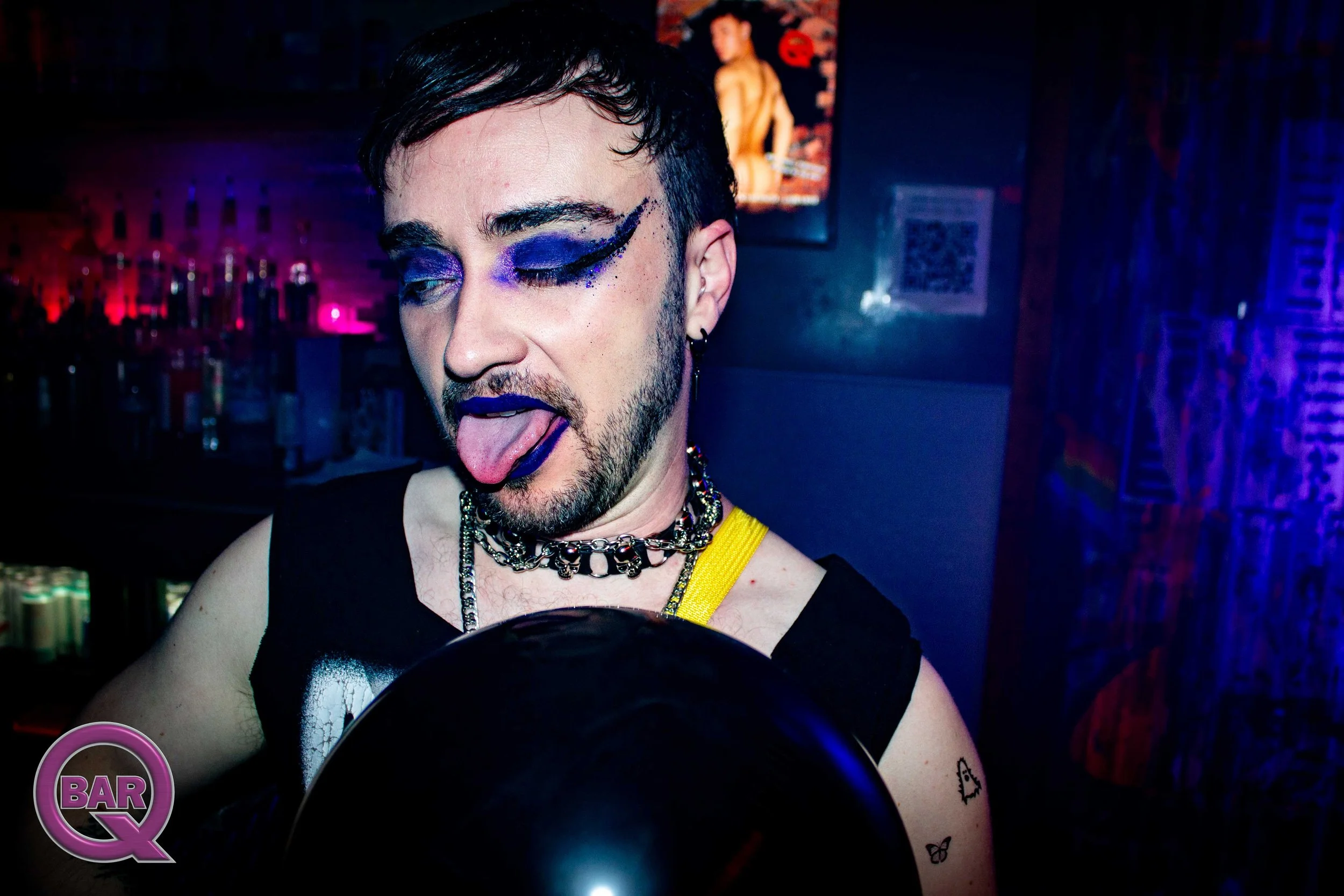 A person with bold purple and black makeup, sticking out their tongue, and dressed in a black tank top with a yellow strap. They are wearing layered chains and earrings. The background appears to be a dimly lit bar or club with colorful decor and pos