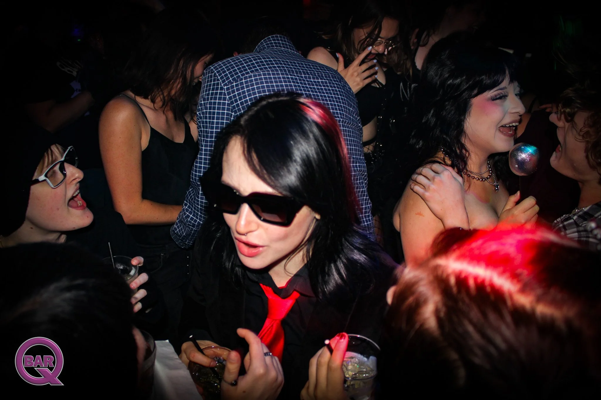 Crowd of people at a lively party or bar, laughing and holding drinks, with a woman in the foreground wearing black sunglasses and a black top.