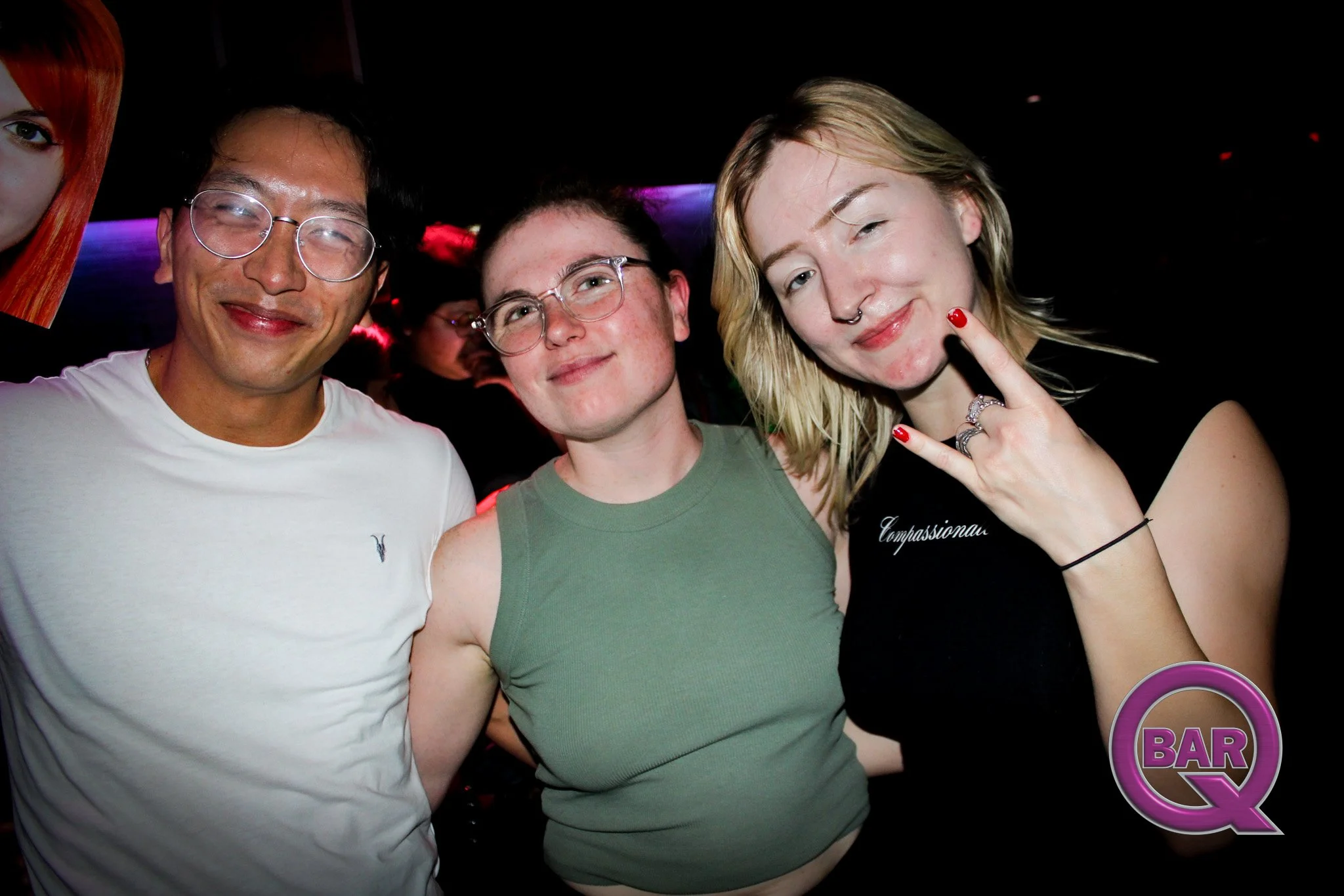 Three people in a bar, smiling and posing for a photo with colorful lighting in the background.