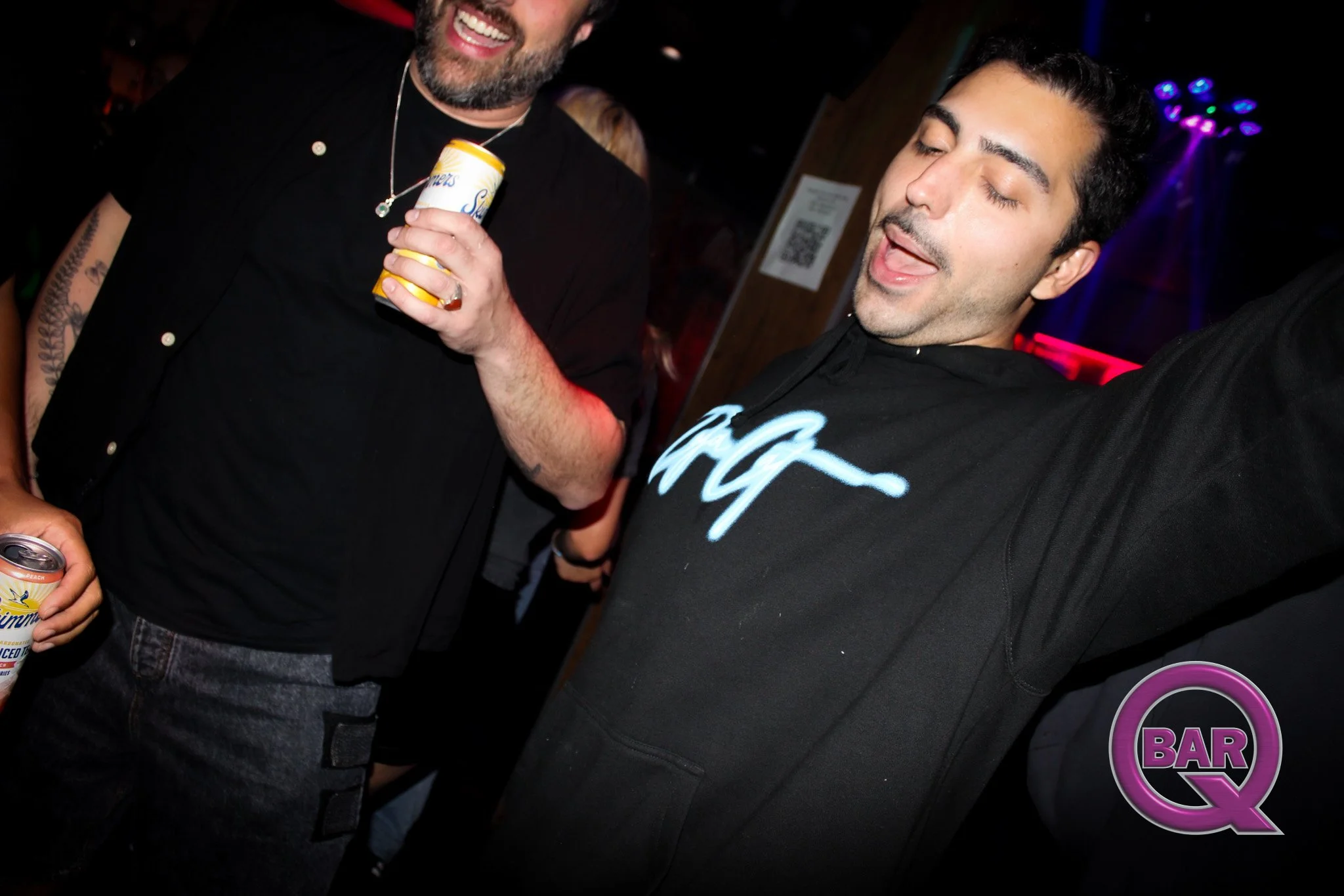 Three people are at a bar, holding drinks. One person on the left is partially visible, holding a can, while the person on the right has their eyes closed and lips slightly parted. The background has colorful lights, and a sign with a QR code is visi