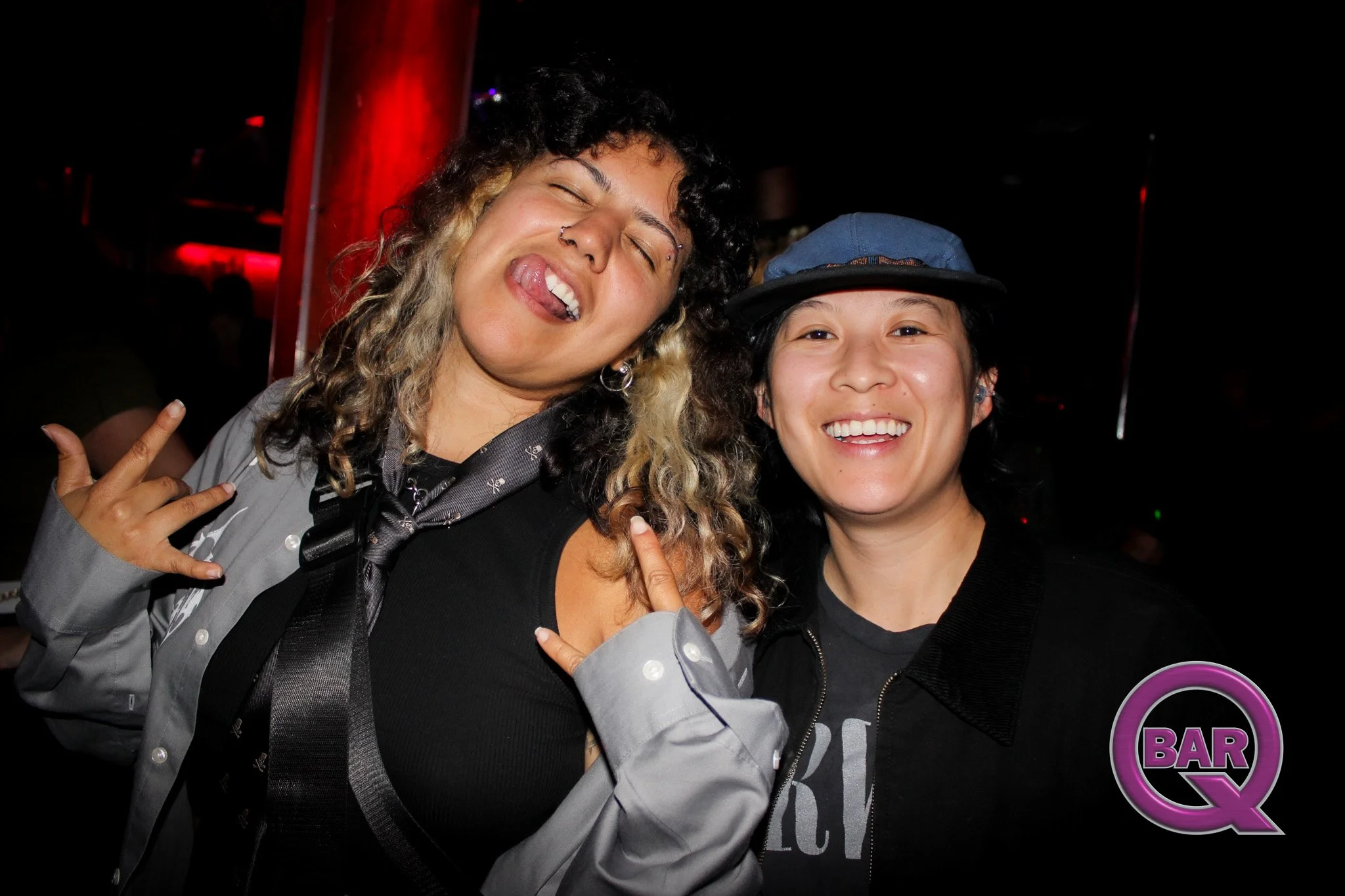 Two smiling women posing together at a bar or club, one with curly hair with blonde tips making rock and roll hand gestures, the other wearing a baseball cap, both showing happiness.