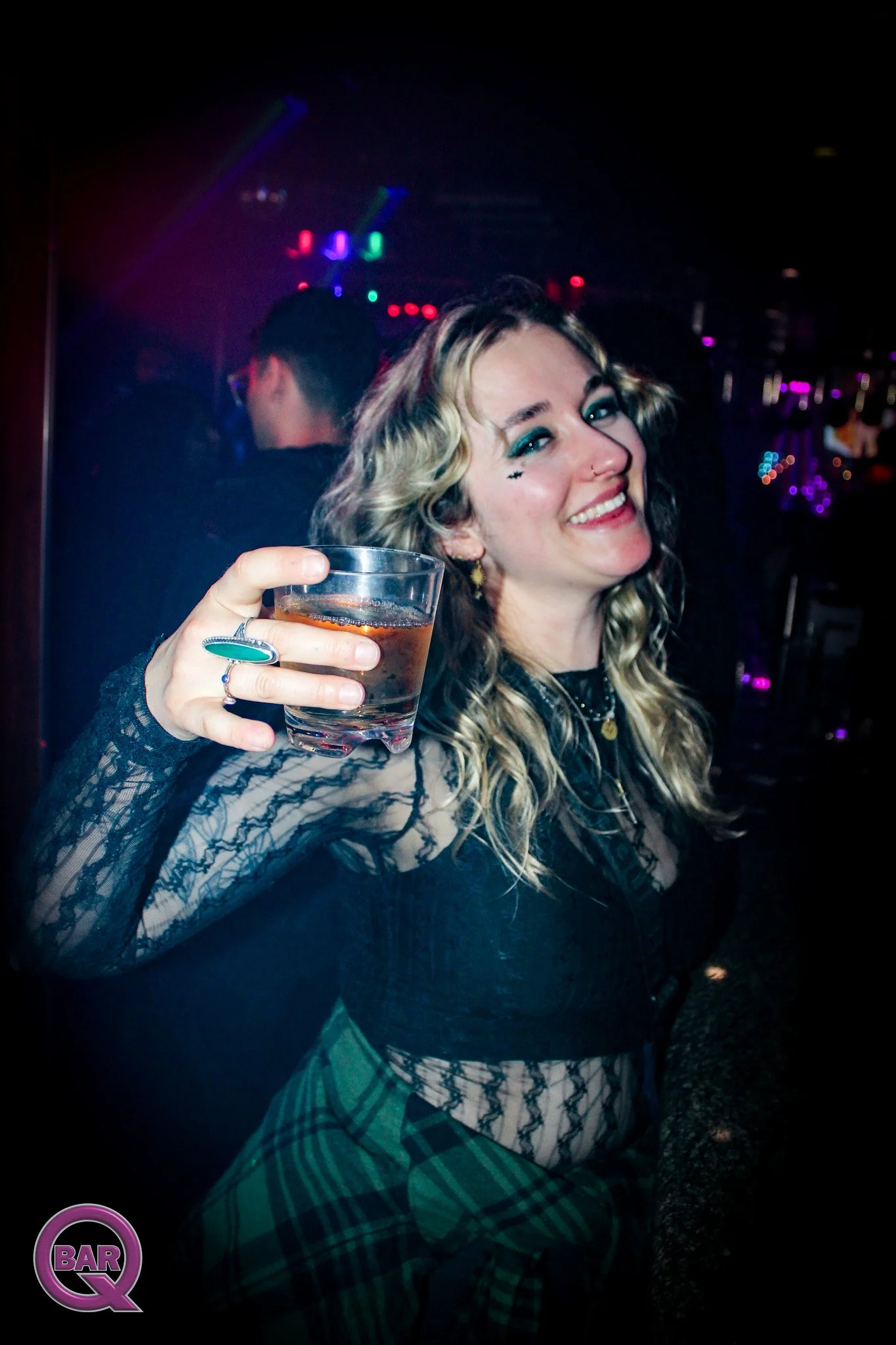 A woman smiling and holding a drink in a dimly lit nightclub or bar with colorful lights in the background.