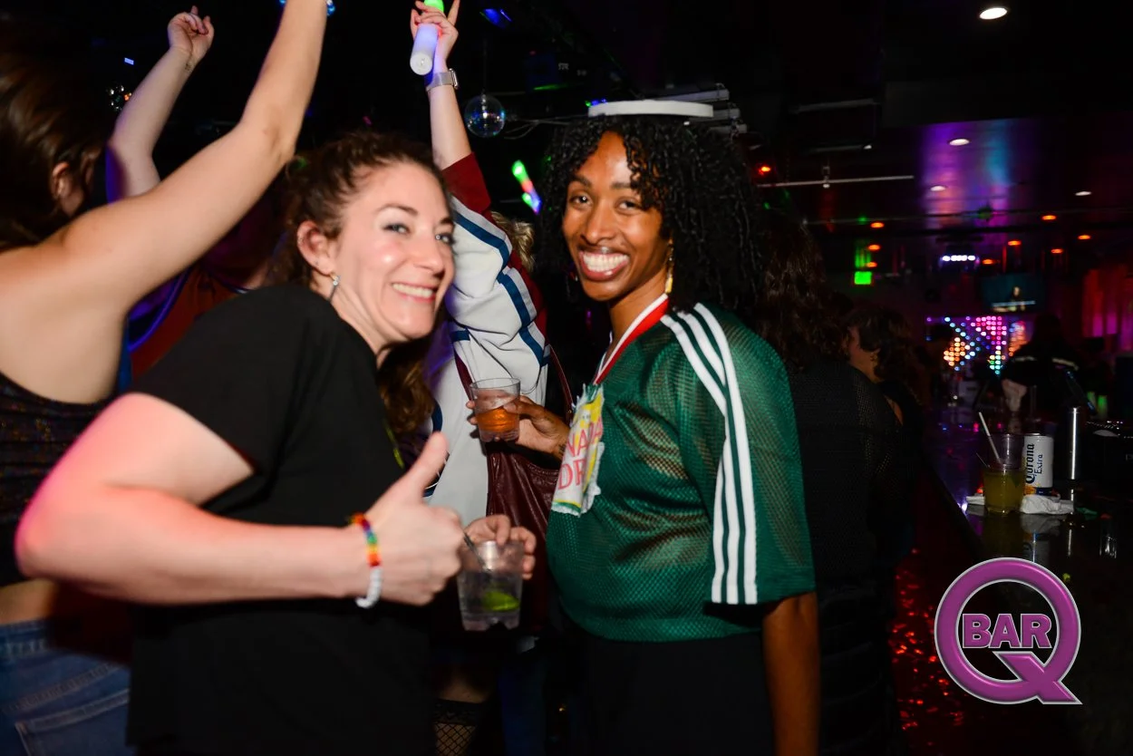 Two women smiling and enjoying drinks at a lively bar or nightclub, surrounded by party-goers. Dark lighting with colorful lights and a 'BAR Q' logo in the corner.