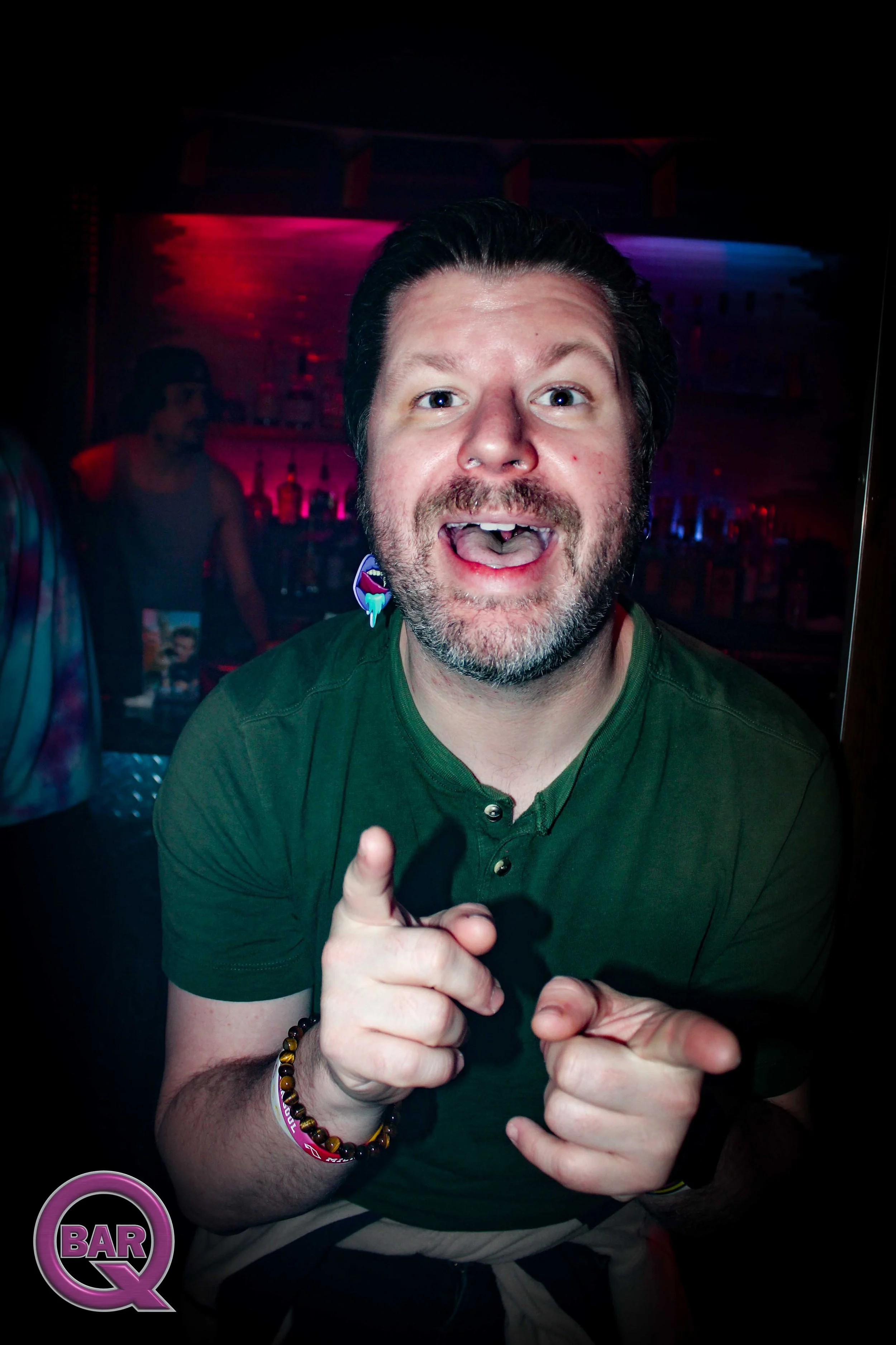 A man at a bar in a dark room with pink and purple lighting, smiling at the camera, wearing a green shirt and various bracelets, pointing at the camera with both hands.