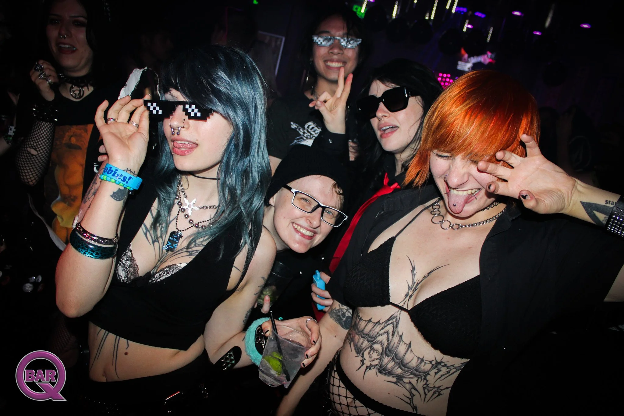 Group of friends at a party, some with colorful hair and tattoos, wearing sunglasses, making peace signs and having fun in a dark, colorful nightclub setting.