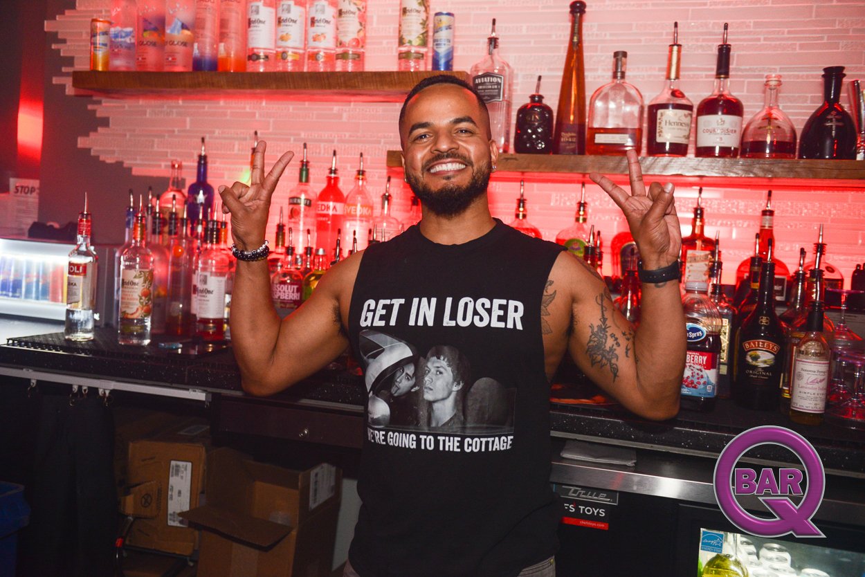 A smiling man with a beard and tattoos wearing a sleeveless shirt, posing with peace signs at a bar with colorful bottles and Red lighting in the background.