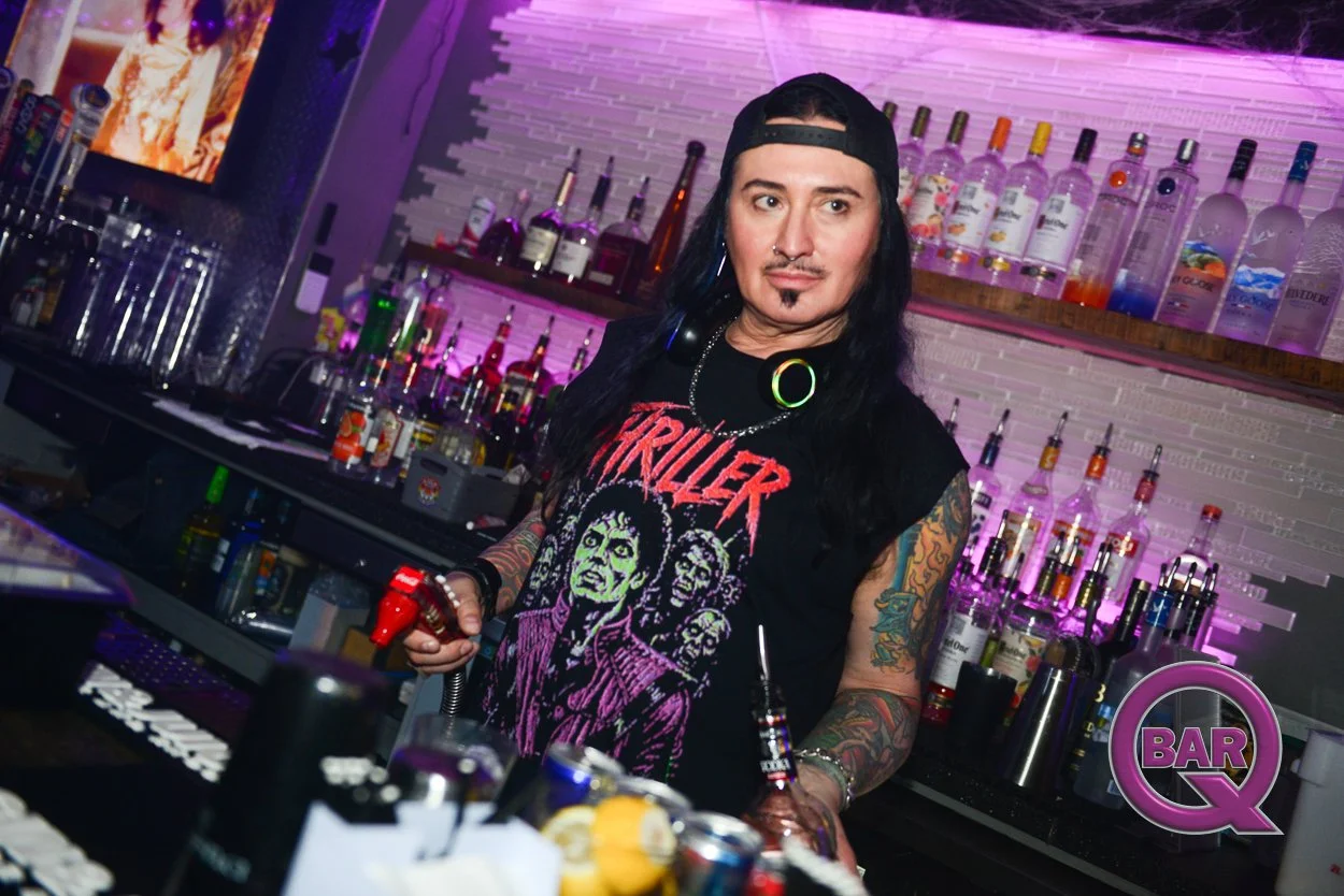 A person with long dark hair, tattoos, and piercings wearing a Black Sabbath "Triller" T-shirt, a backwards baseball cap, and headphones around his neck, standing behind a bar counter filled with bottles of alcohol in a nightclub or bar with colorful