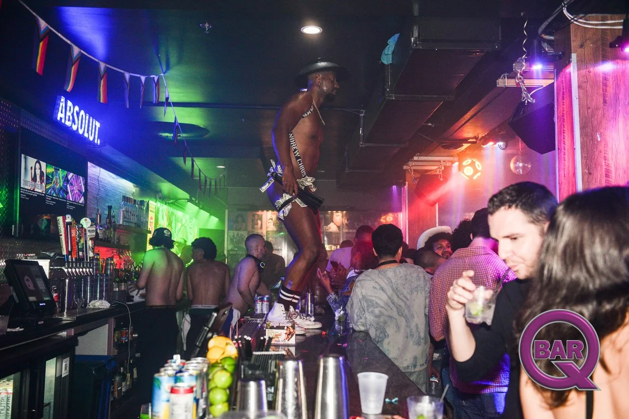 A lively bar scene with a man standing on the bar counter, dancing shirtless in underwear, while holding a gun. The bar is crowded with patrons, some shirtless, and colorful lighting creates a vibrant atmosphere.