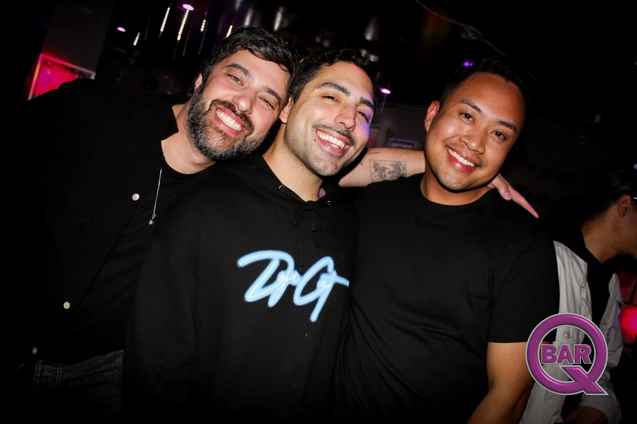 Three smiling men celebrating at a bar or nightclub, standing close together with arms around each other, dark background with pink and purple lighting, and bar signs visible.