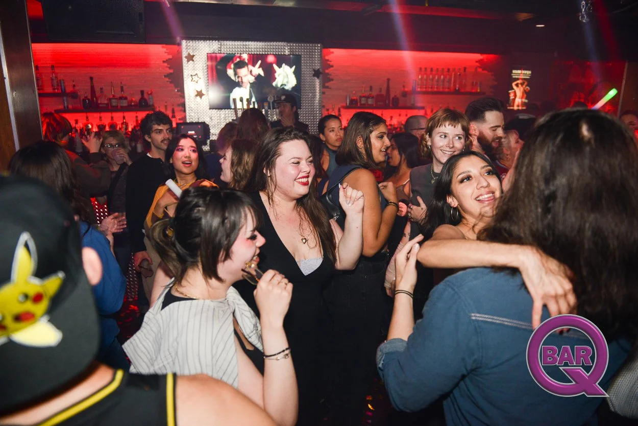 People dancing and socializing in a nightclub with red lighting, a bar in the background, and large screen displaying an image of a man with a microphone. The atmosphere appears lively and festive.