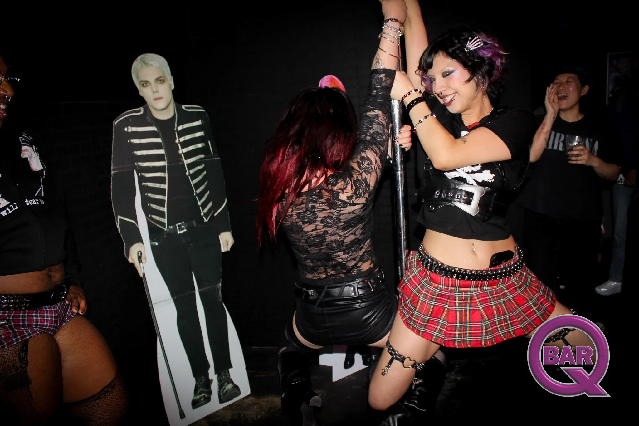 Two women dancing at a bar or nightclub, one with purple and black hair wearing a black top and tartan skirt, the other with red hair wearing black lace top and black leather skirt, a cardboard cutout of a man with platinum blonde hair and black and 