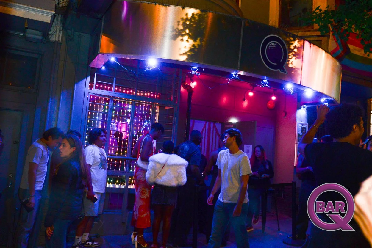 People waiting in line outside a bar with colorful lights and a 'BAR Q' sign, nighttime setting.
