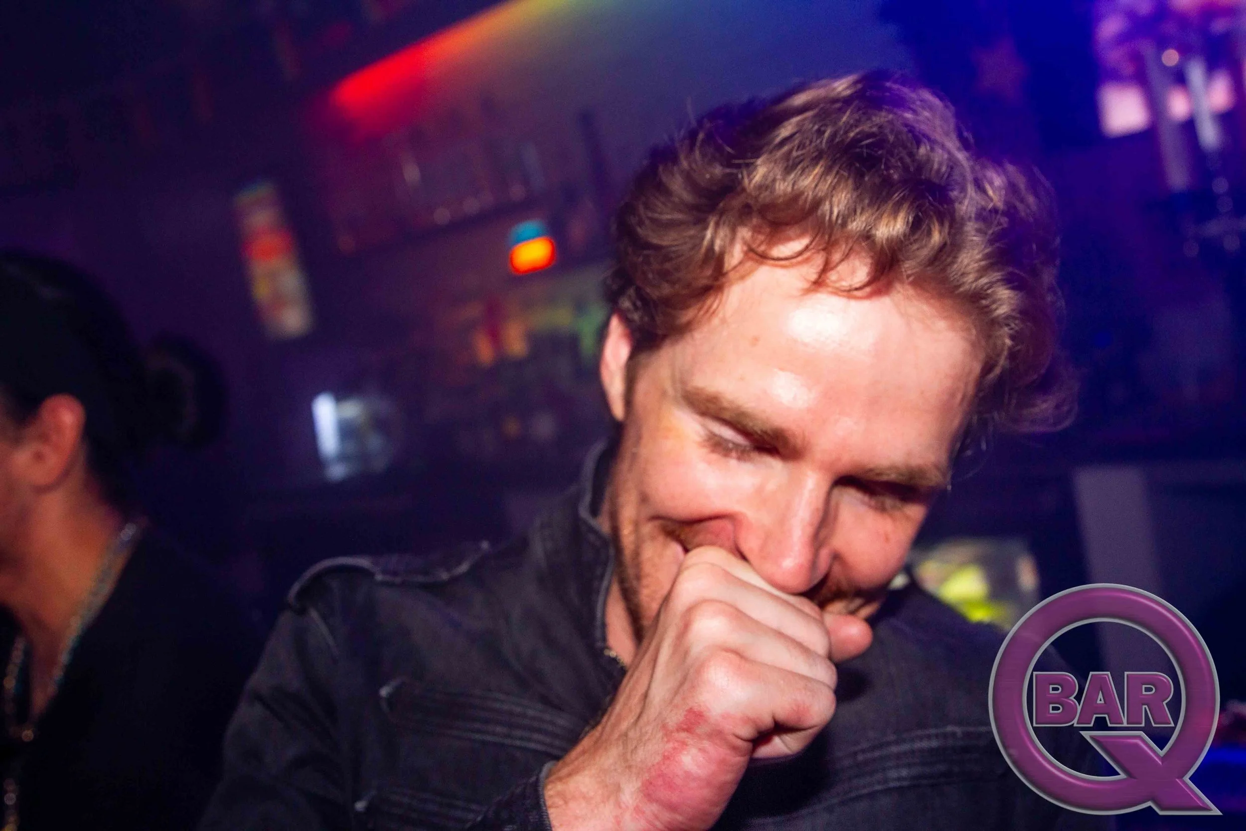 A man with light skin and curly brown hair is smiling while singing into a microphone at a bar or nightclub. The background features colorful lights and a DJ setup.