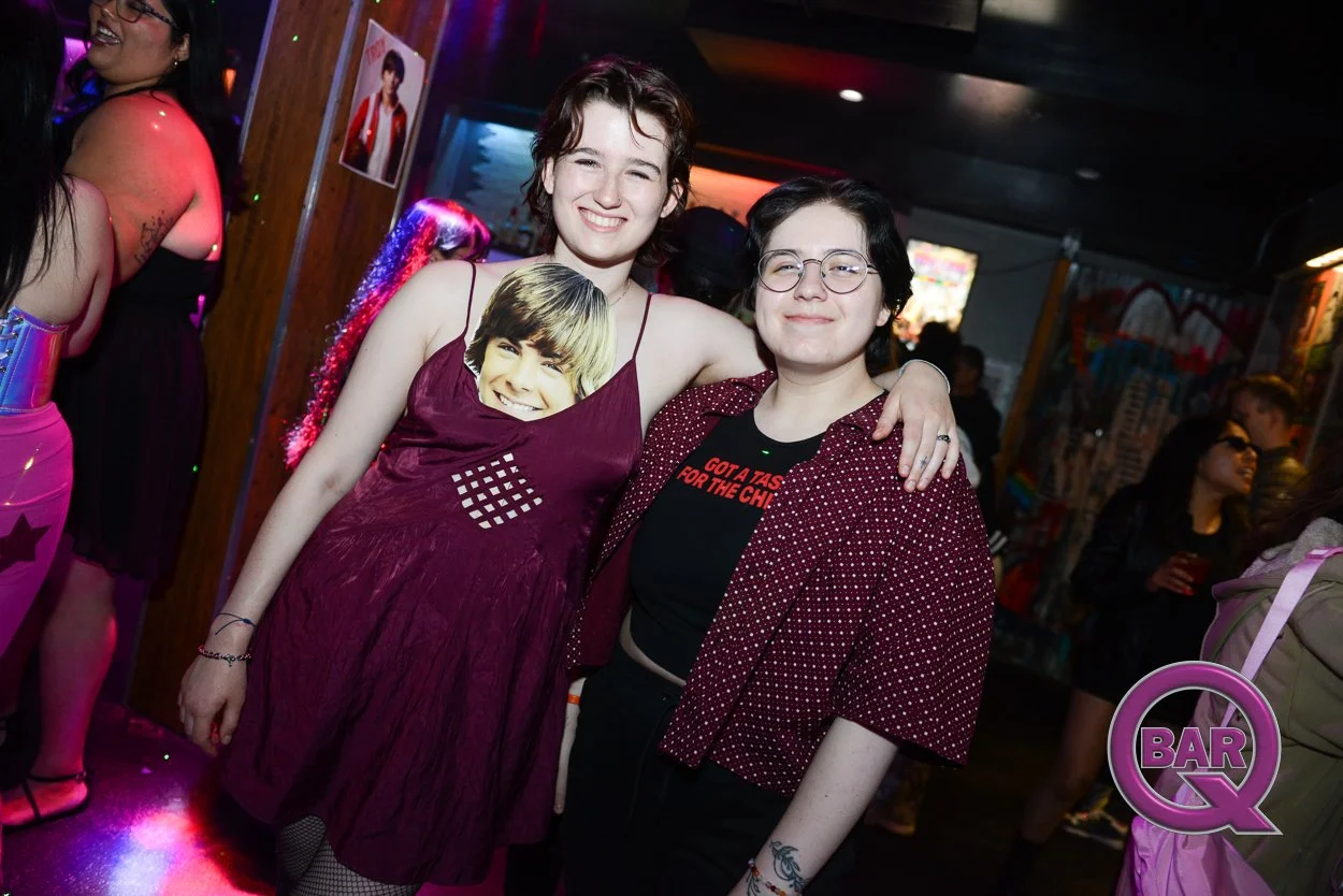 Two young women with short hair and glasses smiling and posing together at a bar or nightclub, with one wearing a dress with a face printed on it and the other in a patterned jacket, surrounded by other people in a colorful, lively setting.