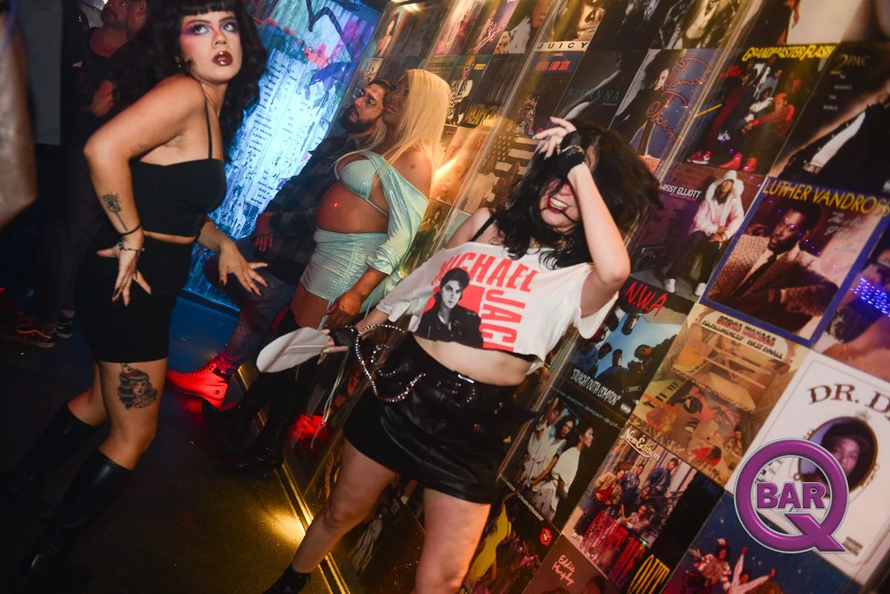 Young woman in a Michael Jackson T-shirt and black leather skirt smiling and holding her head, surrounded by other people in a nightclub decorated with music posters.