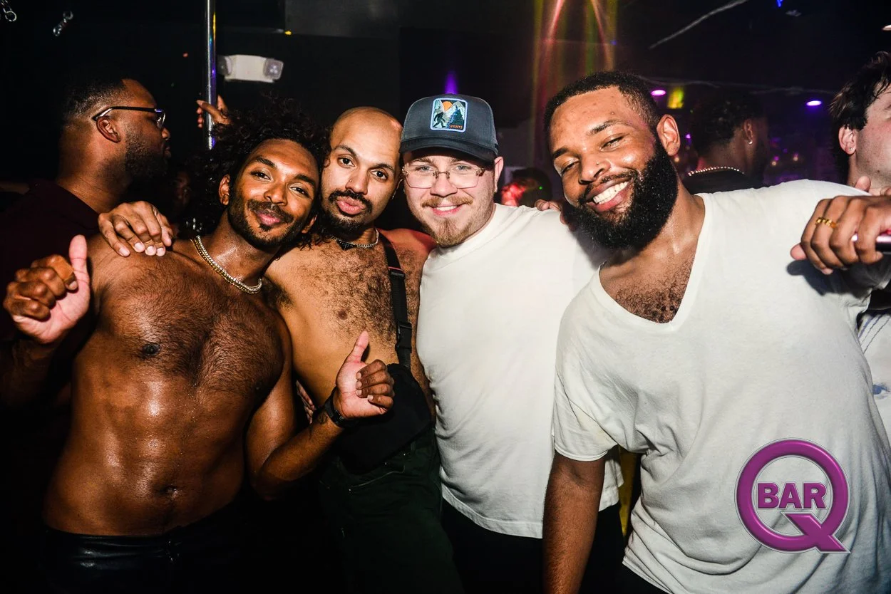 Group of five diverse men smiling and posing together in a nightclub, with low lighting and colorful lights in the background.