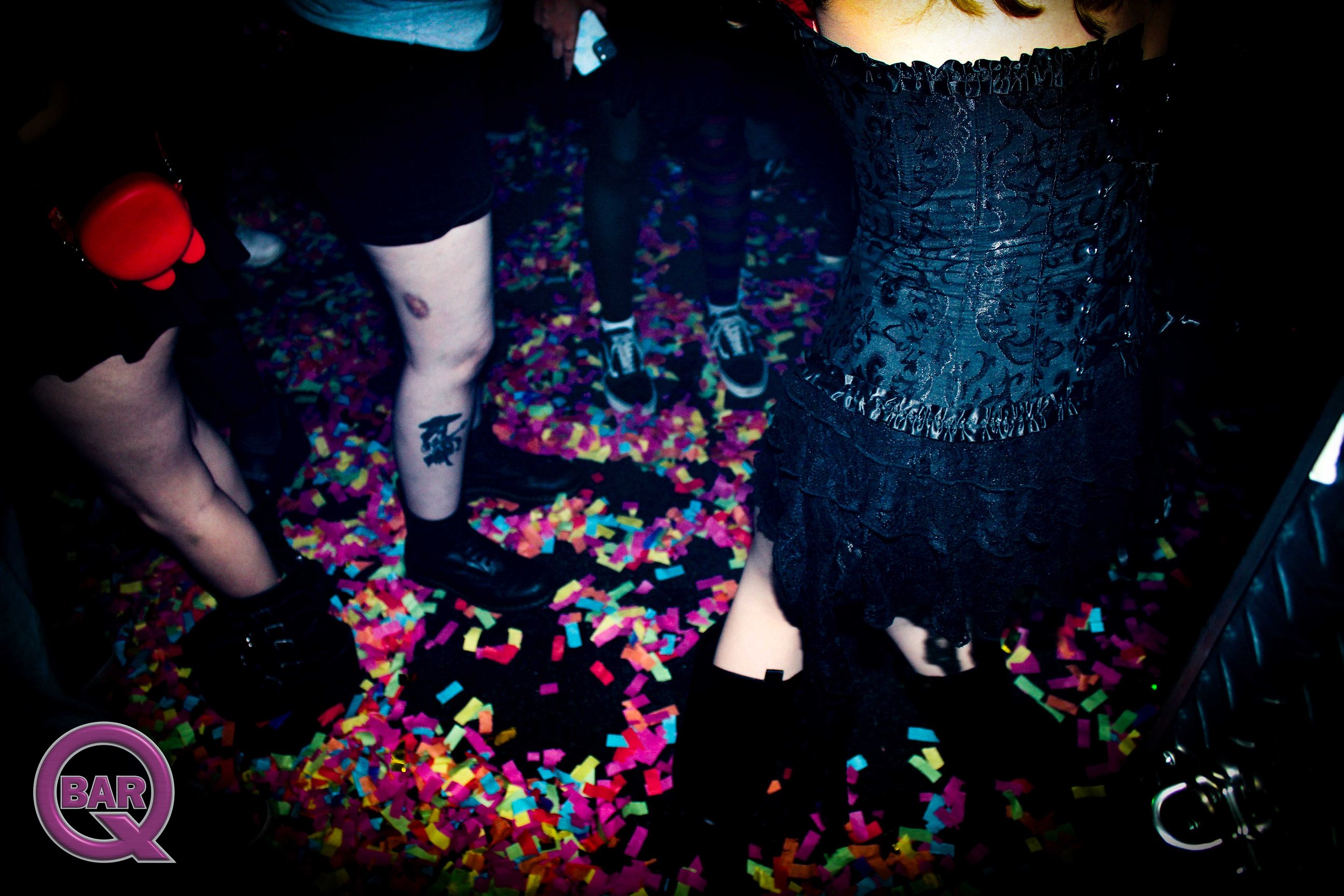 A woman in a black lace dress standing on colorful confetti with other people in the background at a celebration or event.