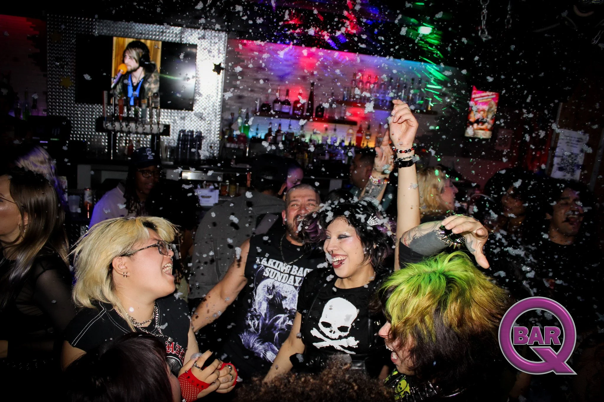 People dancing and celebrating in a bar or nightclub with colorful lights, snow-like confetti, and alcohol bottles in the background.