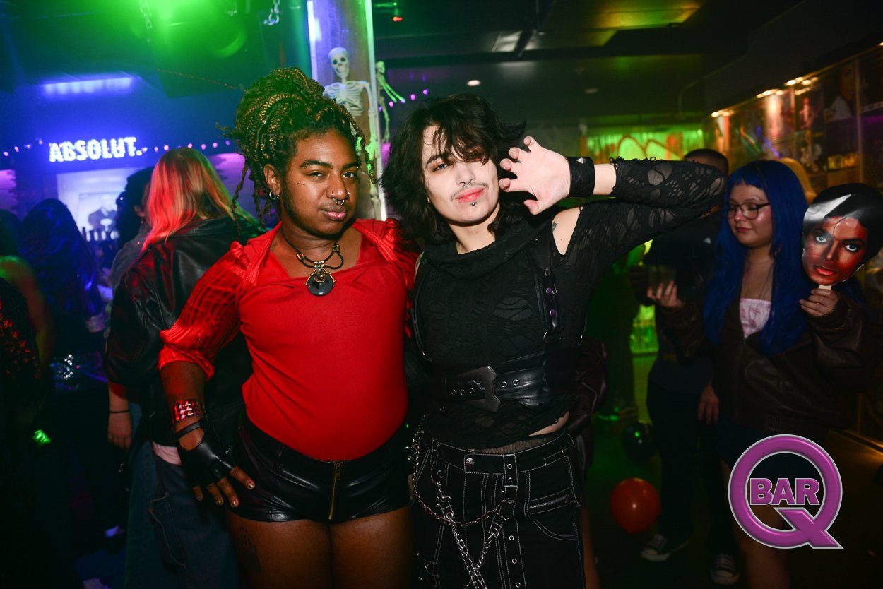 Two women at a nightclub or bar, one with green braided hair and dark skin wearing a red top and black shorts, the other with black hair and light skin wearing a black top. Other people are in the background, some with colorful hair and makeup. Neon 