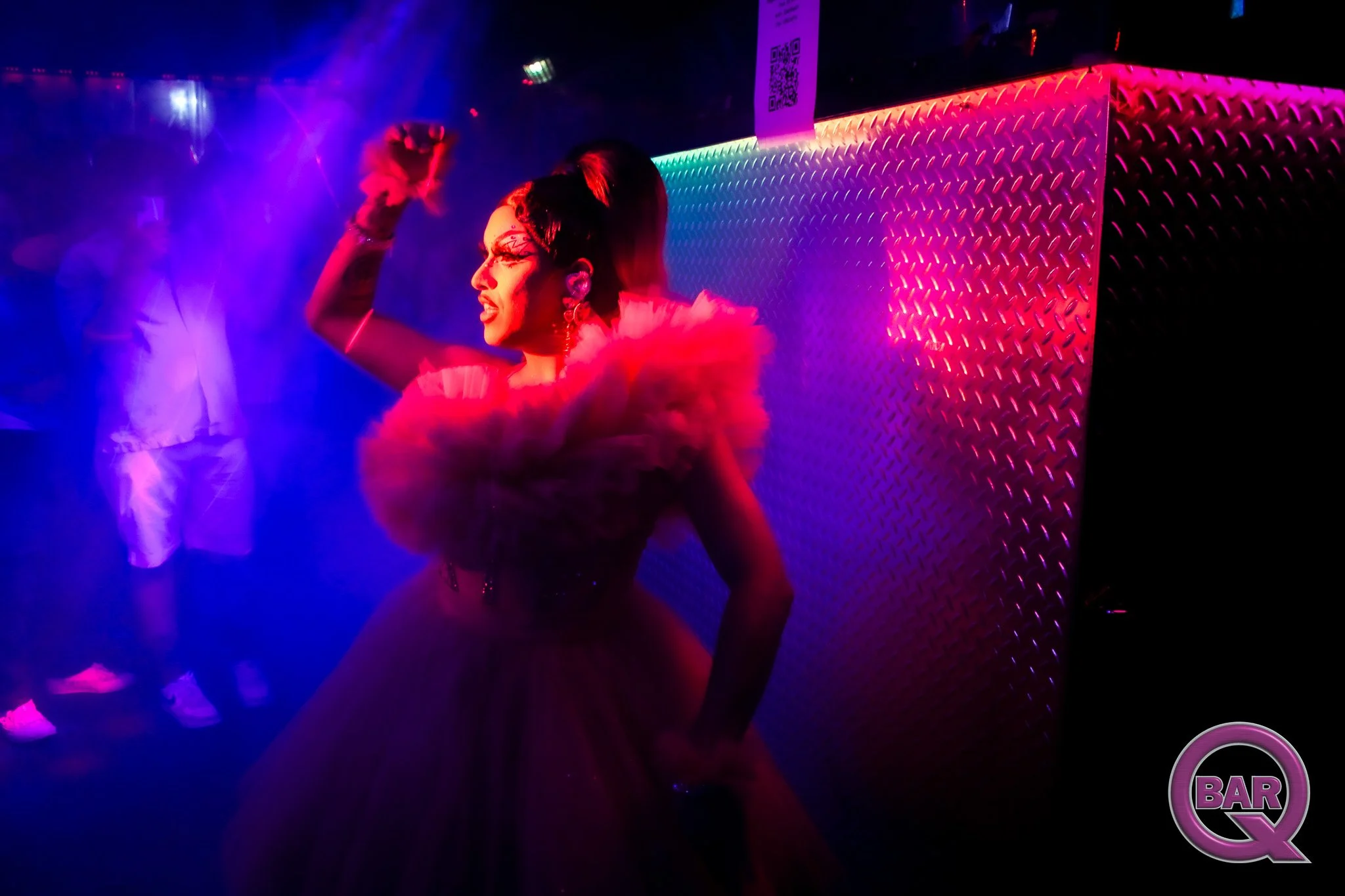 A person dancing in a nightclub with colorful lights, wearing a fuzzy pink outfit, with a bar sign visible in the background.