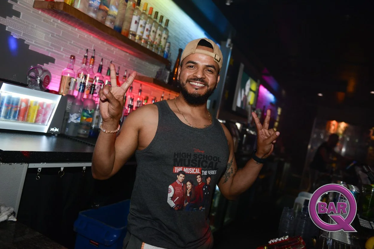 Man smiling and making peace signs at a bar, wearing a tank top and a cap backwards.