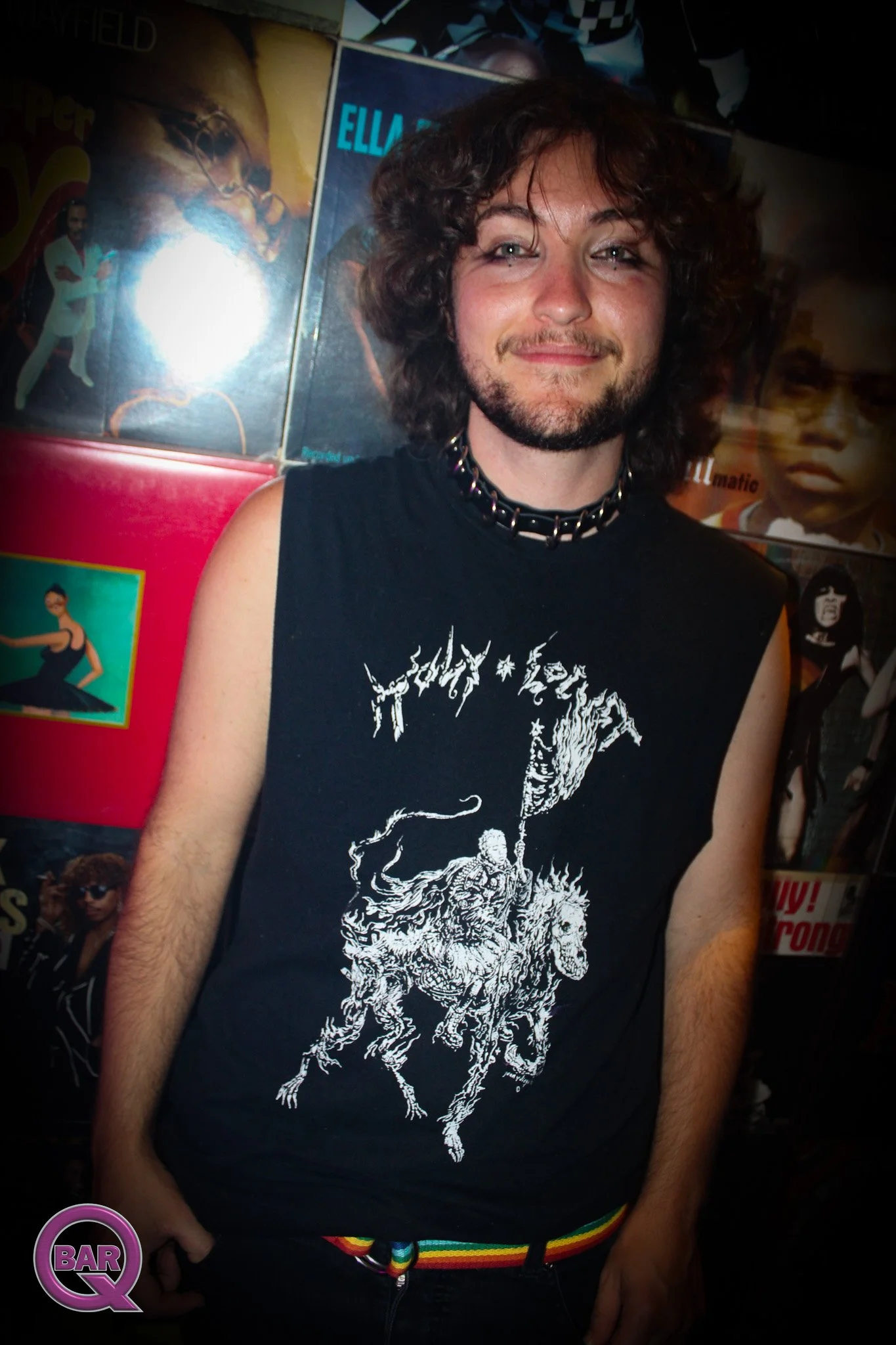 A person with curly hair and a beard wearing a black sleeveless shirt with a white graphic of a lion on it and a rainbow striped belt, standing in front of a wall decorated with various posters, including one of a man with sunglasses and another of a